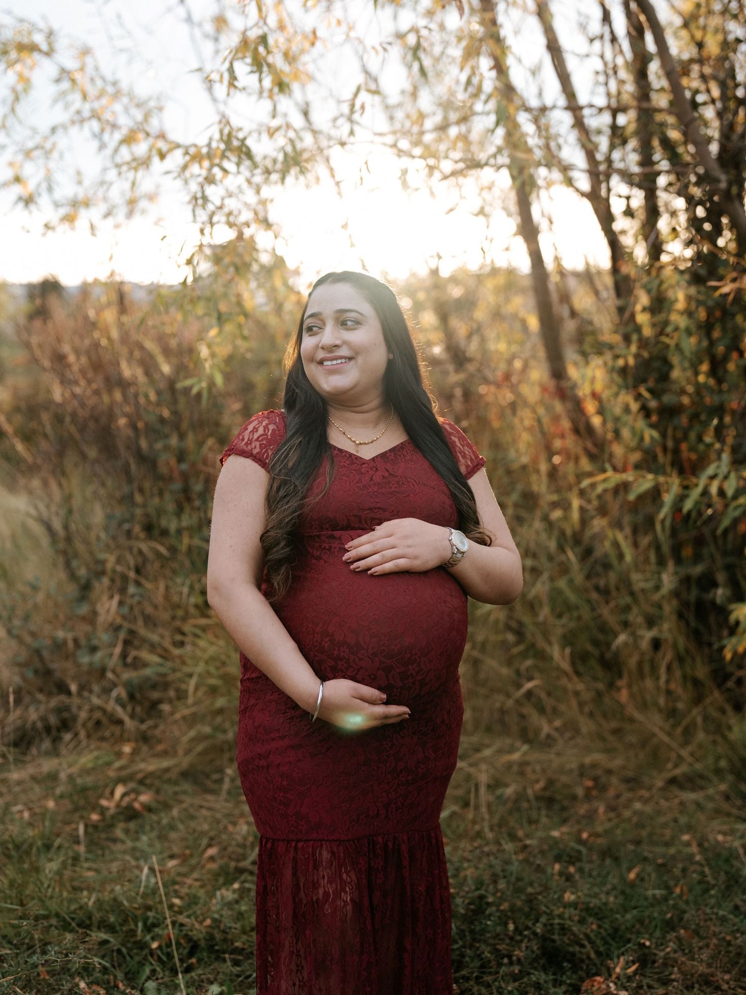 A maternity portrait in a burgundy dress against golden sunset foliage.