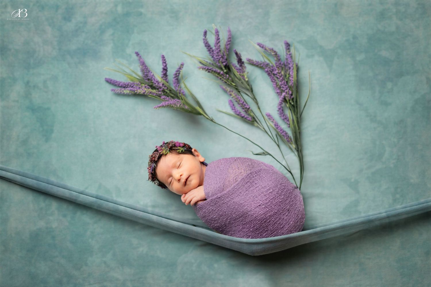 Swaddled baby in purple wrap sleeping on curved surface with lavender sprigs.