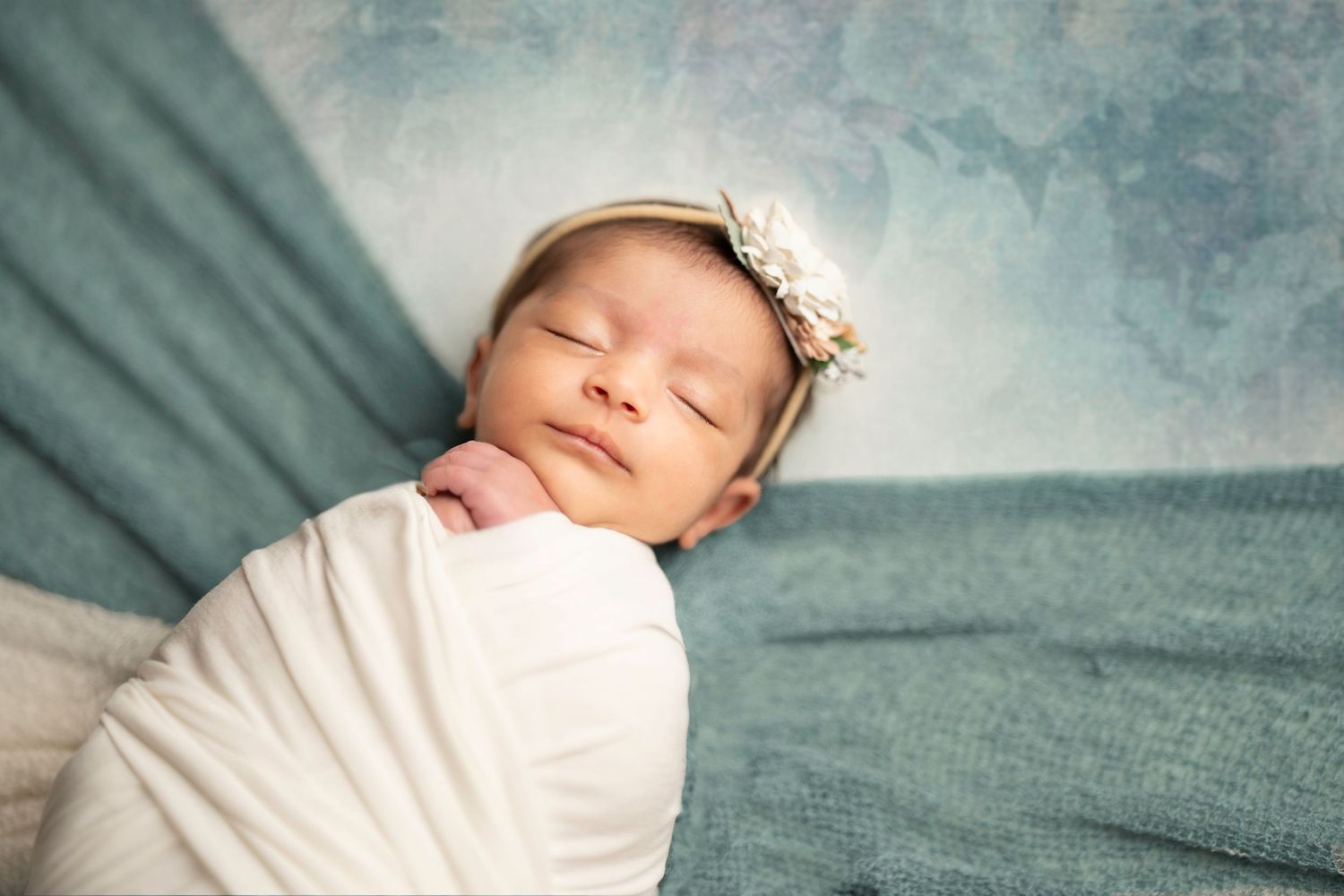 Sleeping newborn in white wrap against teal backdrop with delicate headpiece.