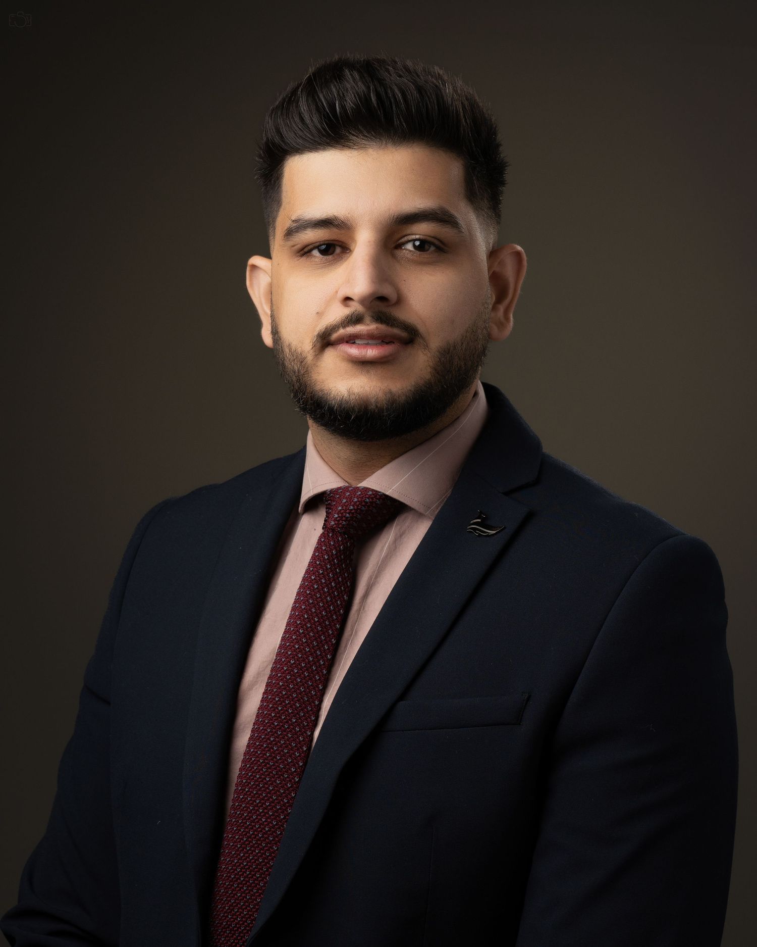 Professional headshot of a man in navy suit and burgundy tie against dark background.