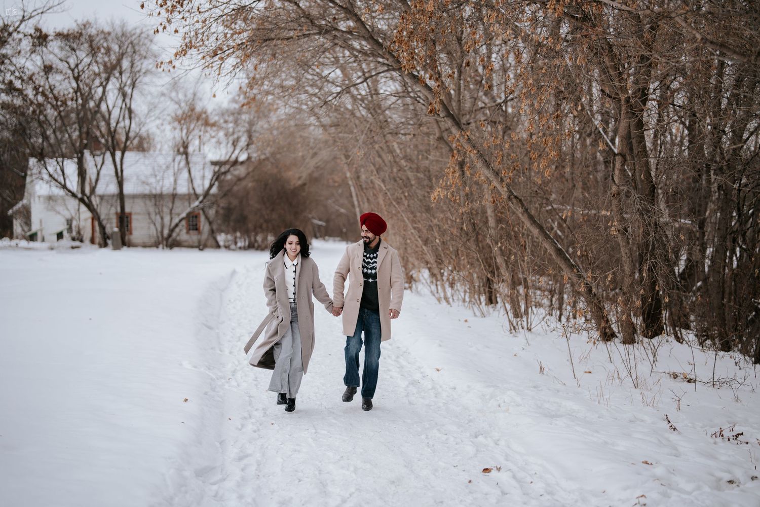 Romantic couple walking hand in hand through a snow-covered path surrounded by bare winter trees.