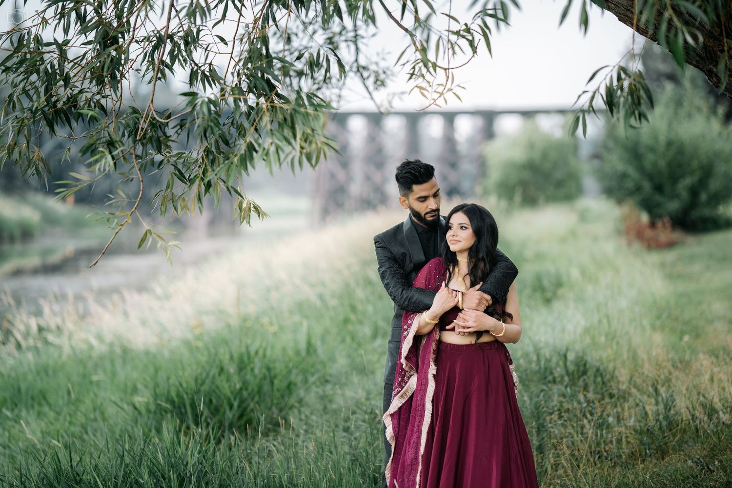 Couple embraces under willow trees in romantic outdoor setting.
