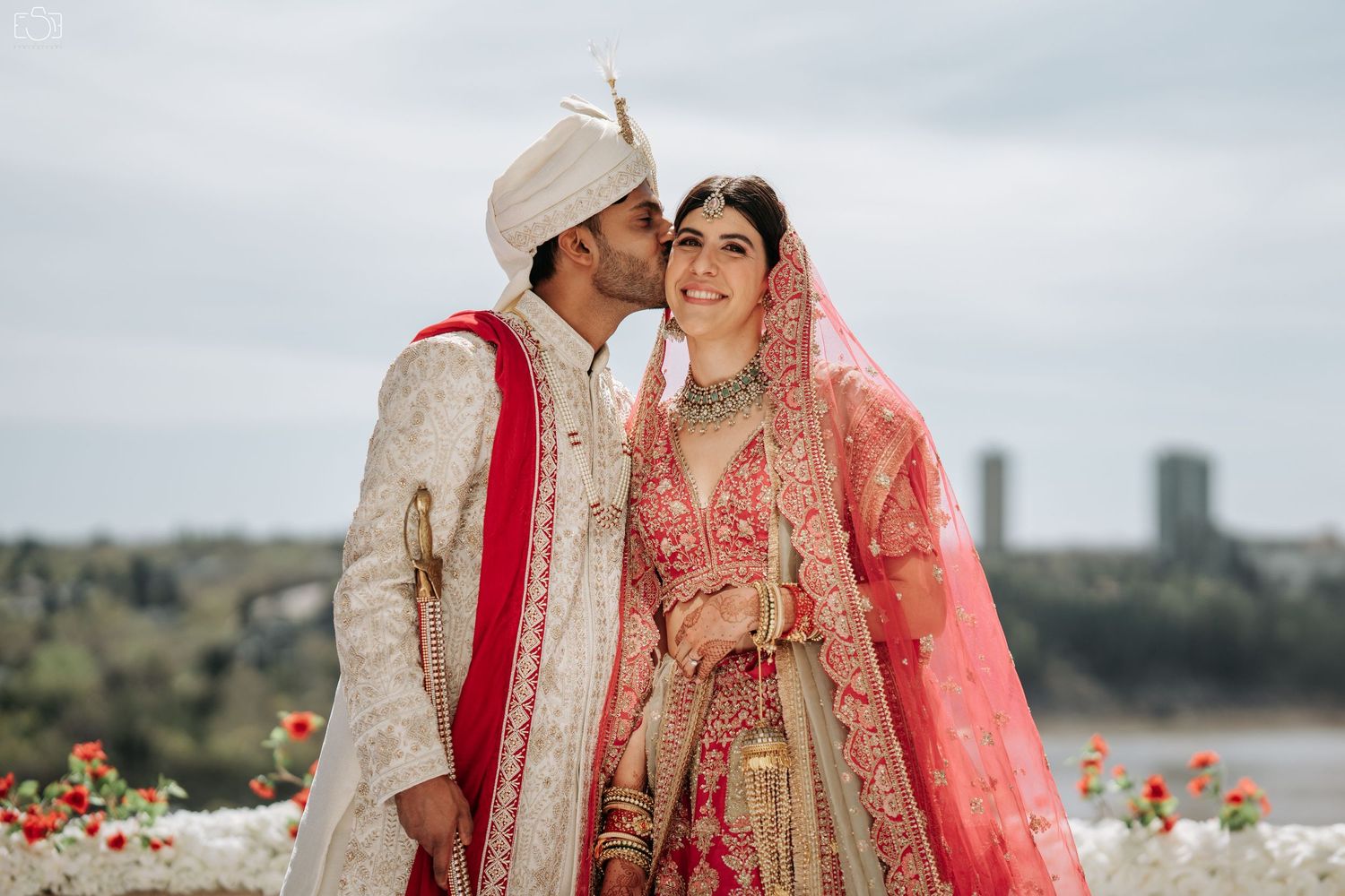 Traditional Indian wedding couple in elaborate red and cream outfits against cloudy sky.