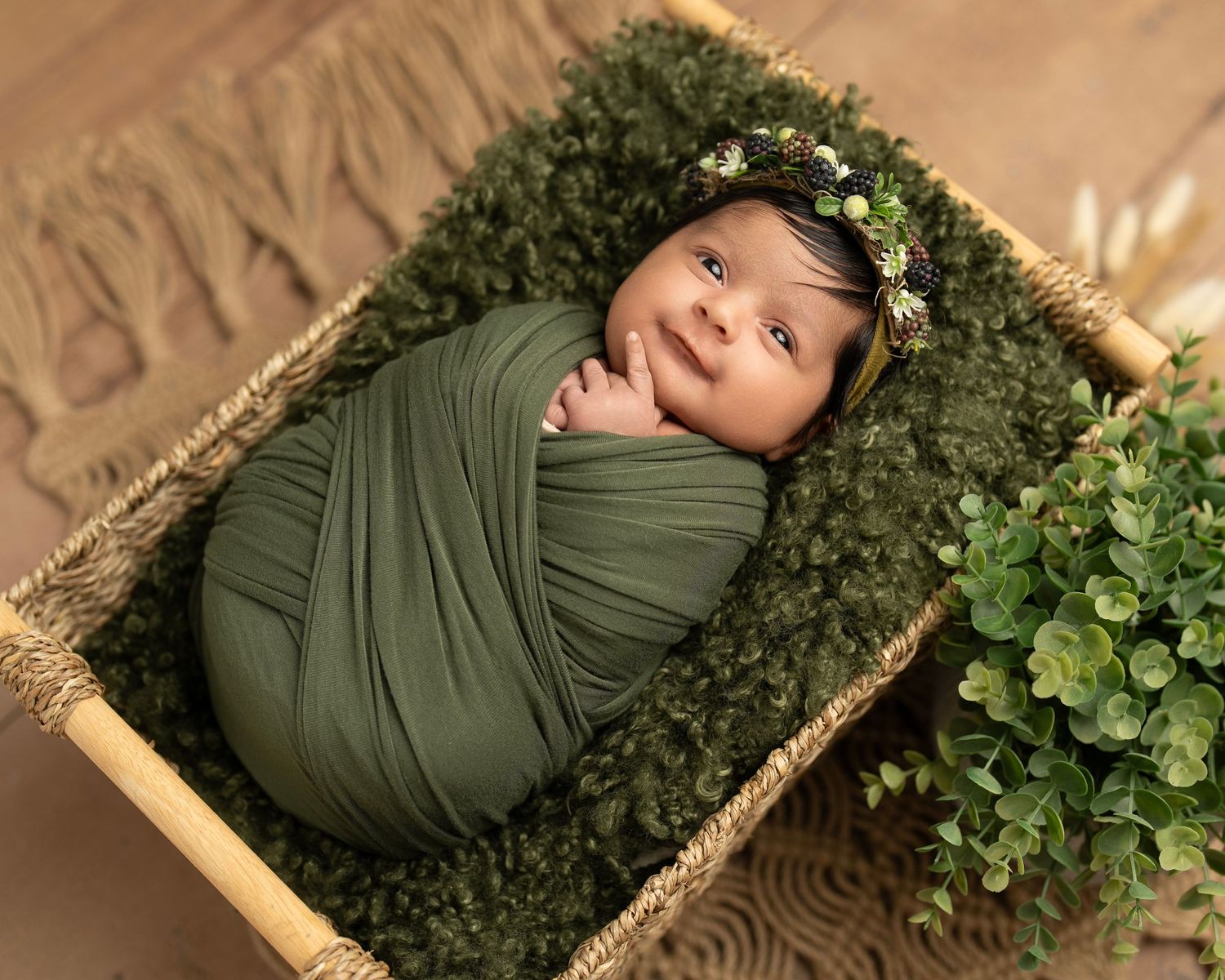Wrapped newborn in olive green swaddle rests peacefully in basket with floral crown.