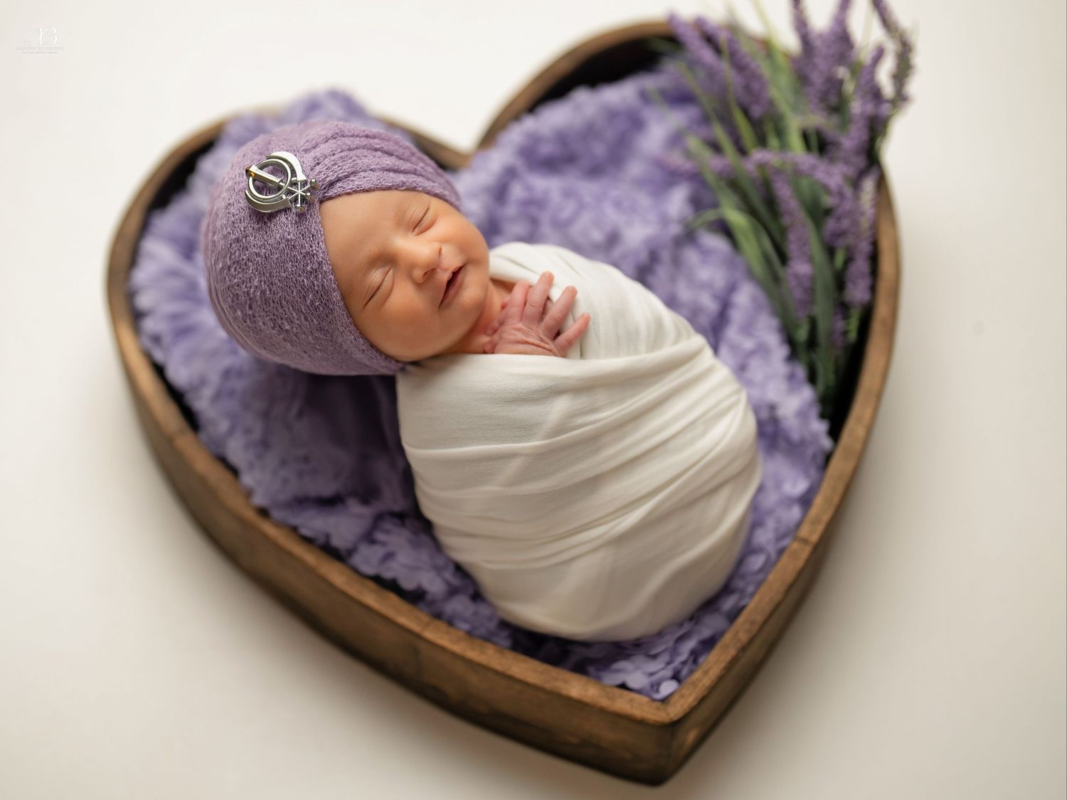 Sleeping newborn in white wrap and purple bonnet nestled in heart-shaped wooden bowl.