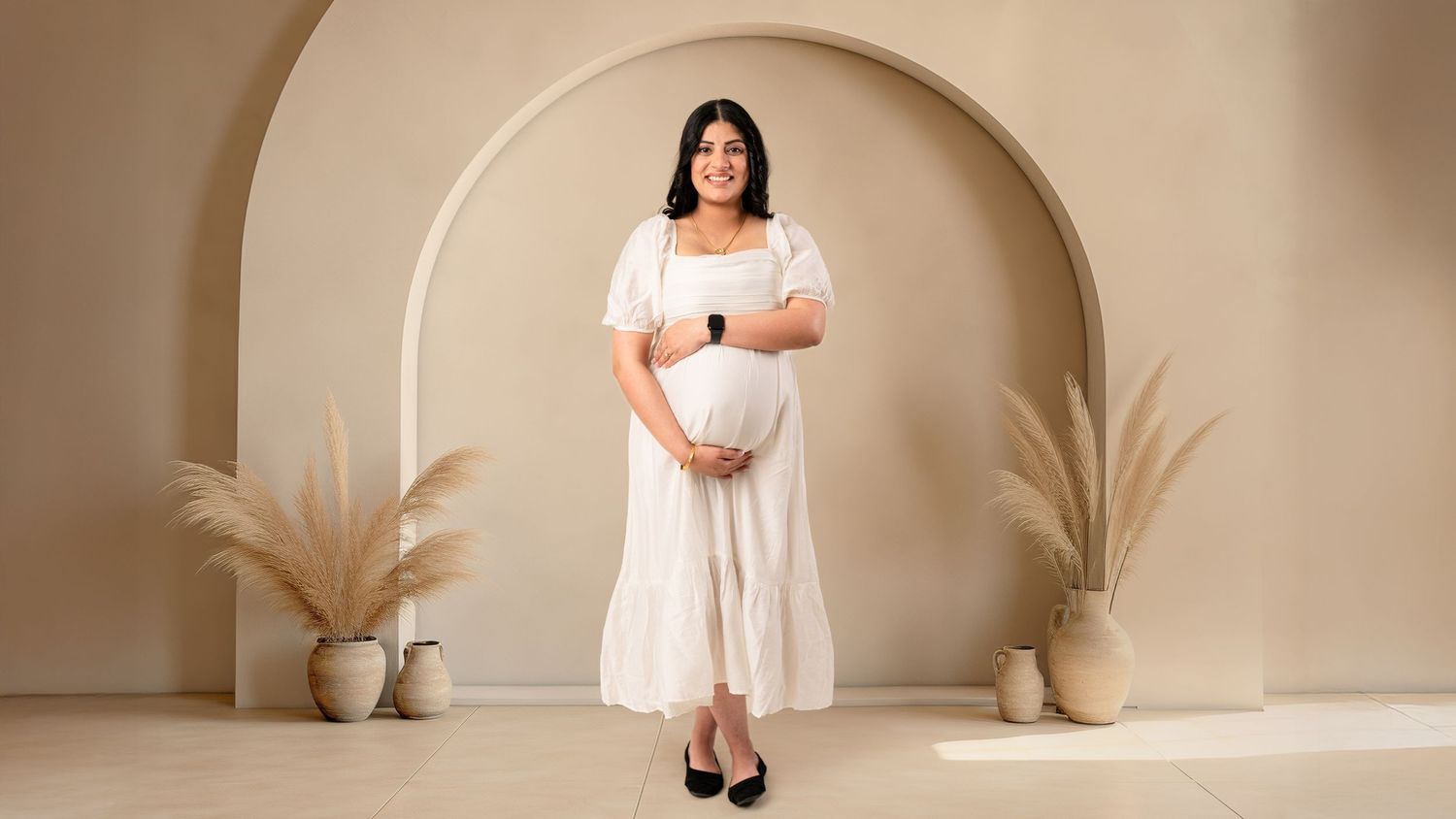 An elegant maternity portrait in a white dress against a beige arched background.