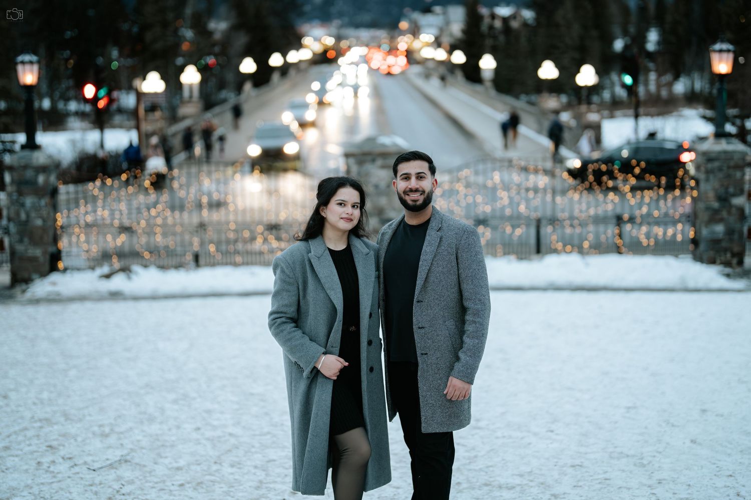 Stylish couple poses on snowy path lined with twinkling lights at dusk.