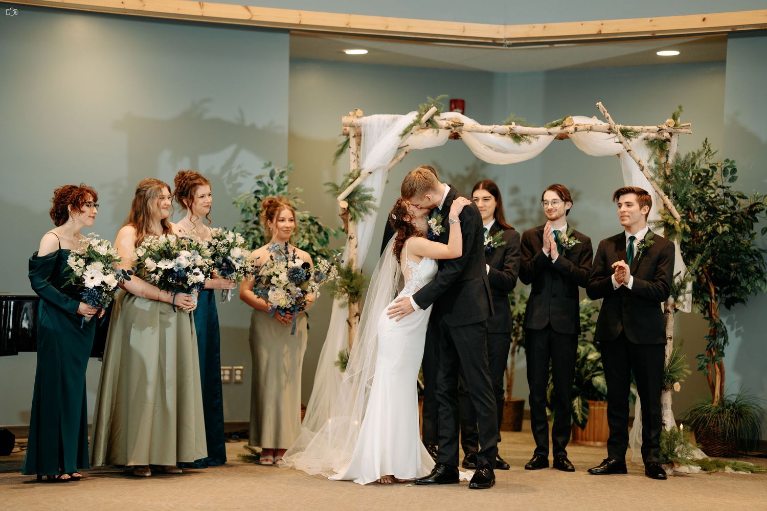 Newlyweds share first kiss under floral arch while wedding party looks on.