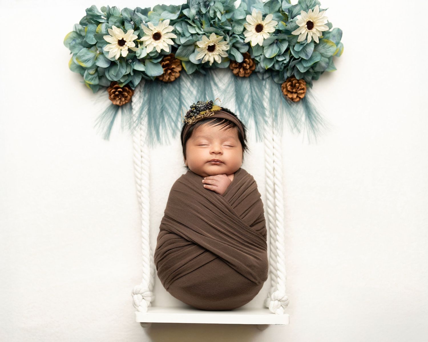 Sleeping newborn swaddled in brown wrap rests beneath floral arch on white swing.