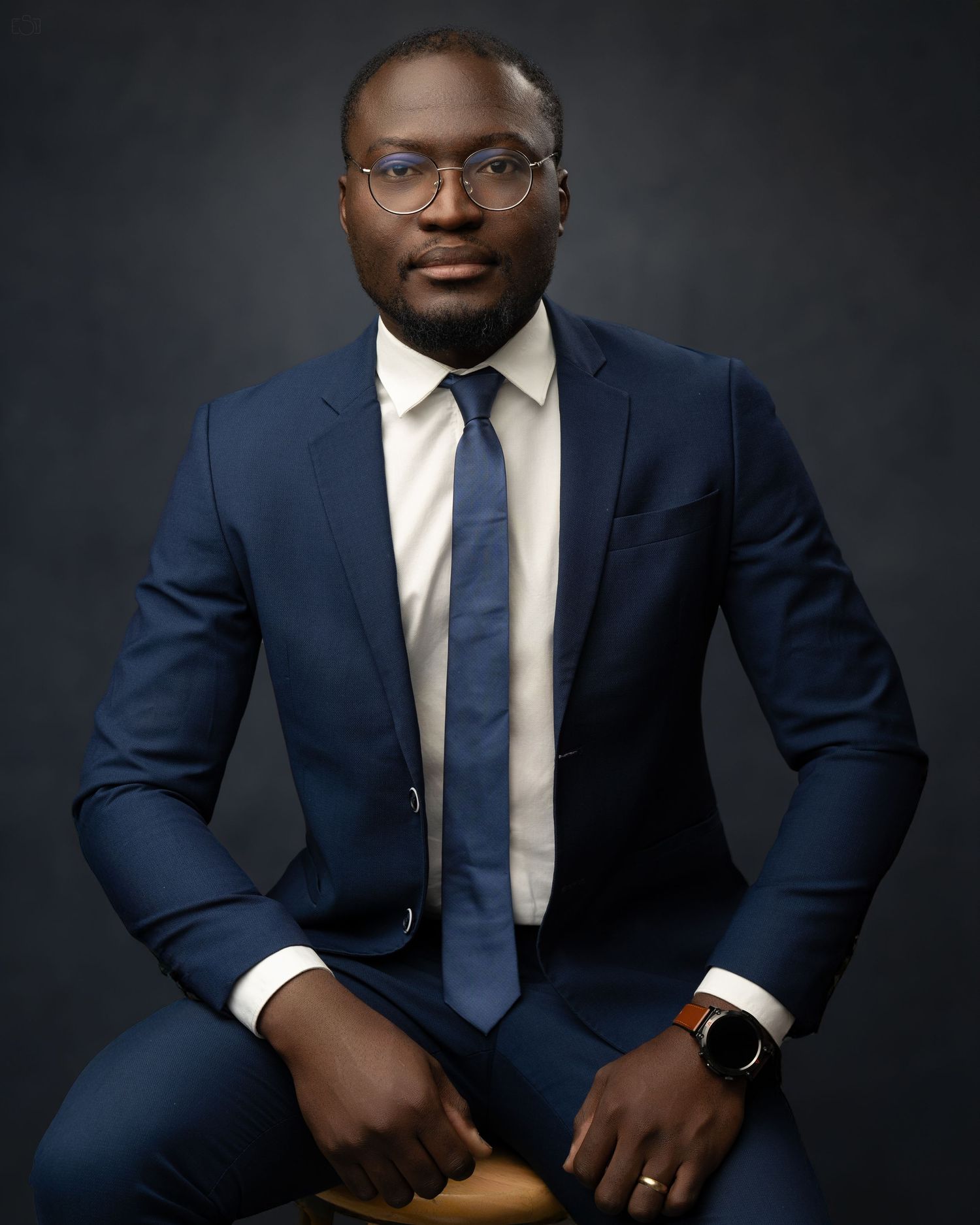 Professional headshot against dark background featuring navy suit and blue tie.