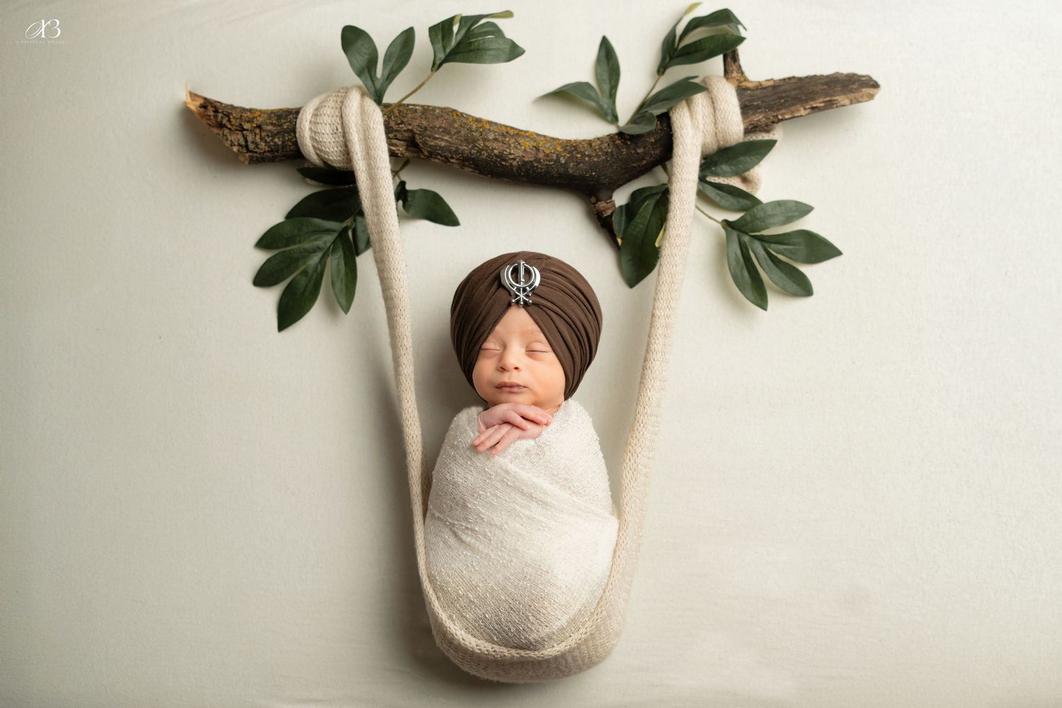 Sleeping newborn swaddled in white fabric hanging from wooden branch with green leaves.