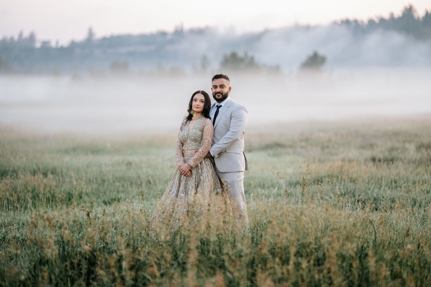 A couple stands together in a misty meadow field during golden hour as fog rolls across the landscape.