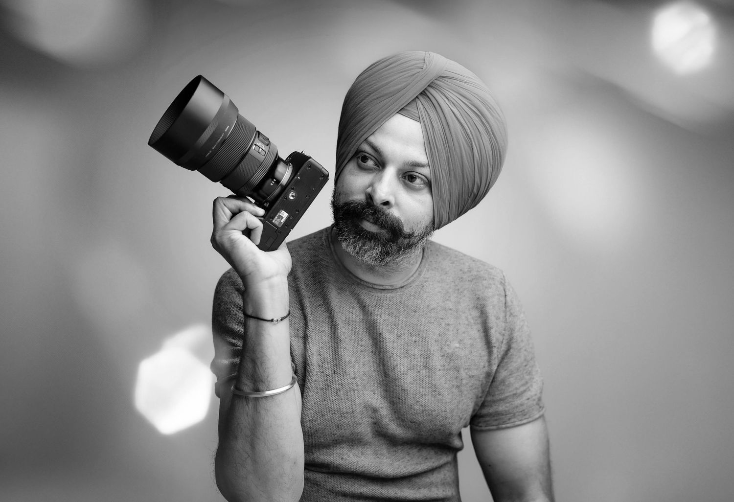 A photographer wearing a turban holds up a camera with a telephoto lens in a black and white portrait.