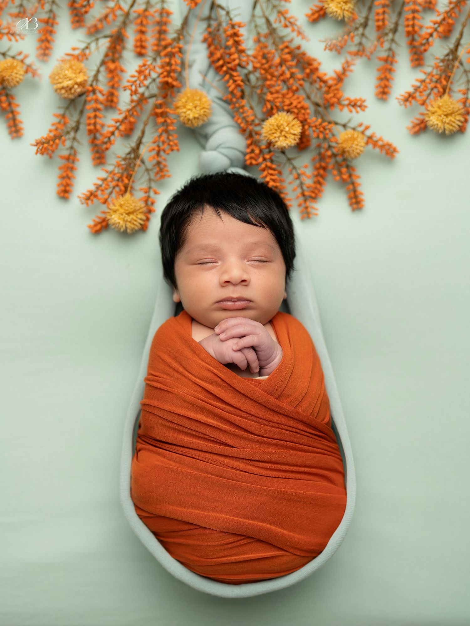 Newborn sleeping peacefully in orange wrap under hanging orange floral branches.