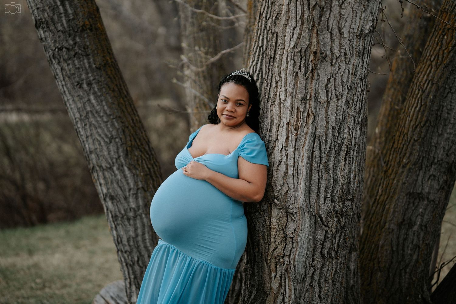 Pregnant woman in blue dress smiling while posing between two trees.