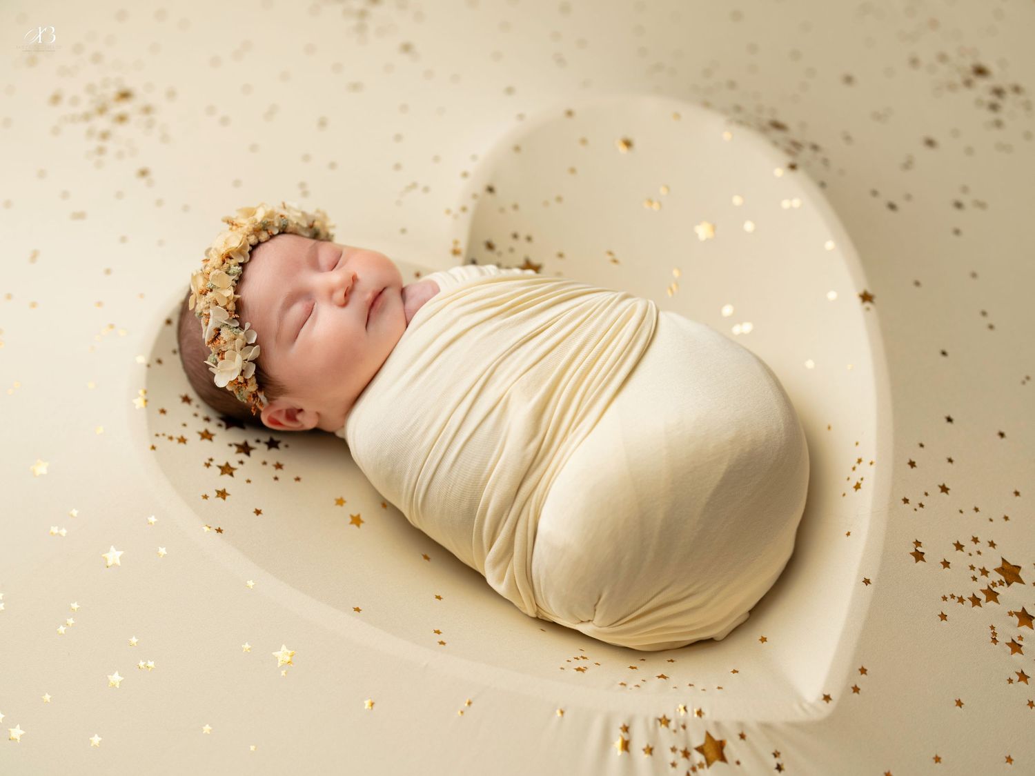Peaceful newborn in cream wrap with gold crown sleeps amid sparkly backdrop.