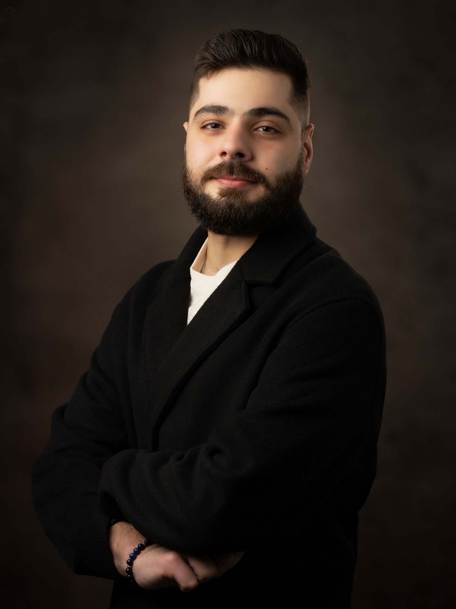Studio portrait of a person in a black coat with a well-groomed beard against a dark backdrop.