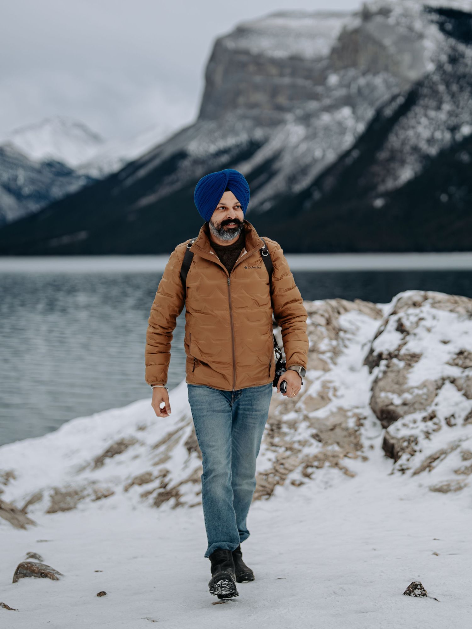 Person in a blue turban and brown jacket hiking along a snowy lakeshore with majestic mountain peaks in the background.