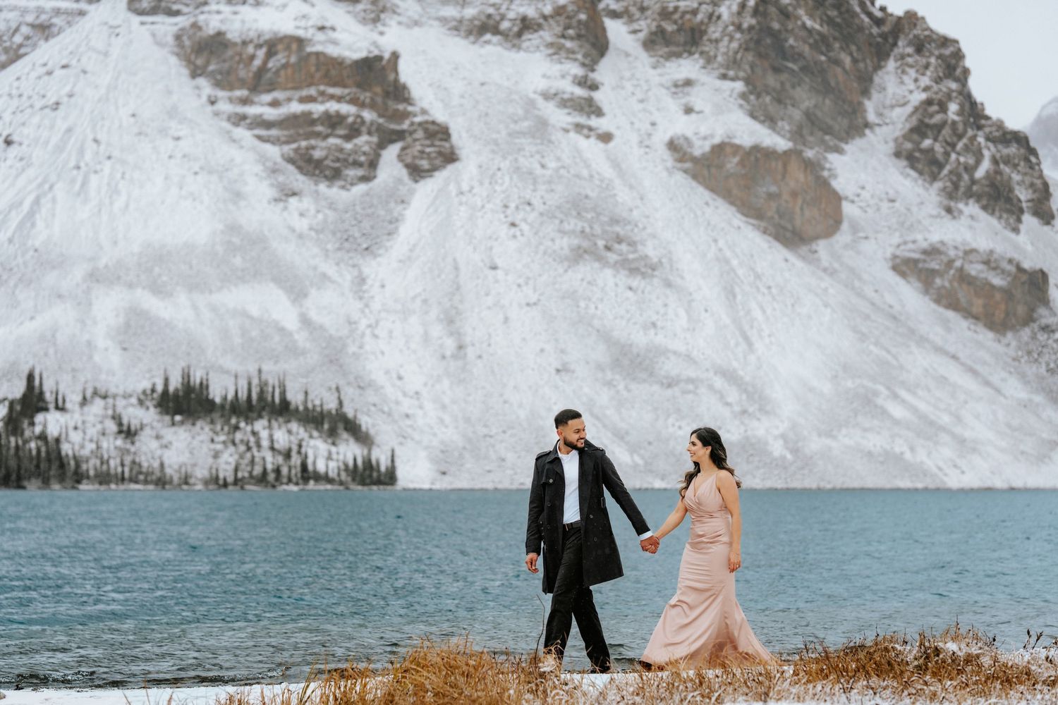 A couple stands on the shore of a turquoise lake surrounded by snow-capped mountains in a dramatic winter landscape.