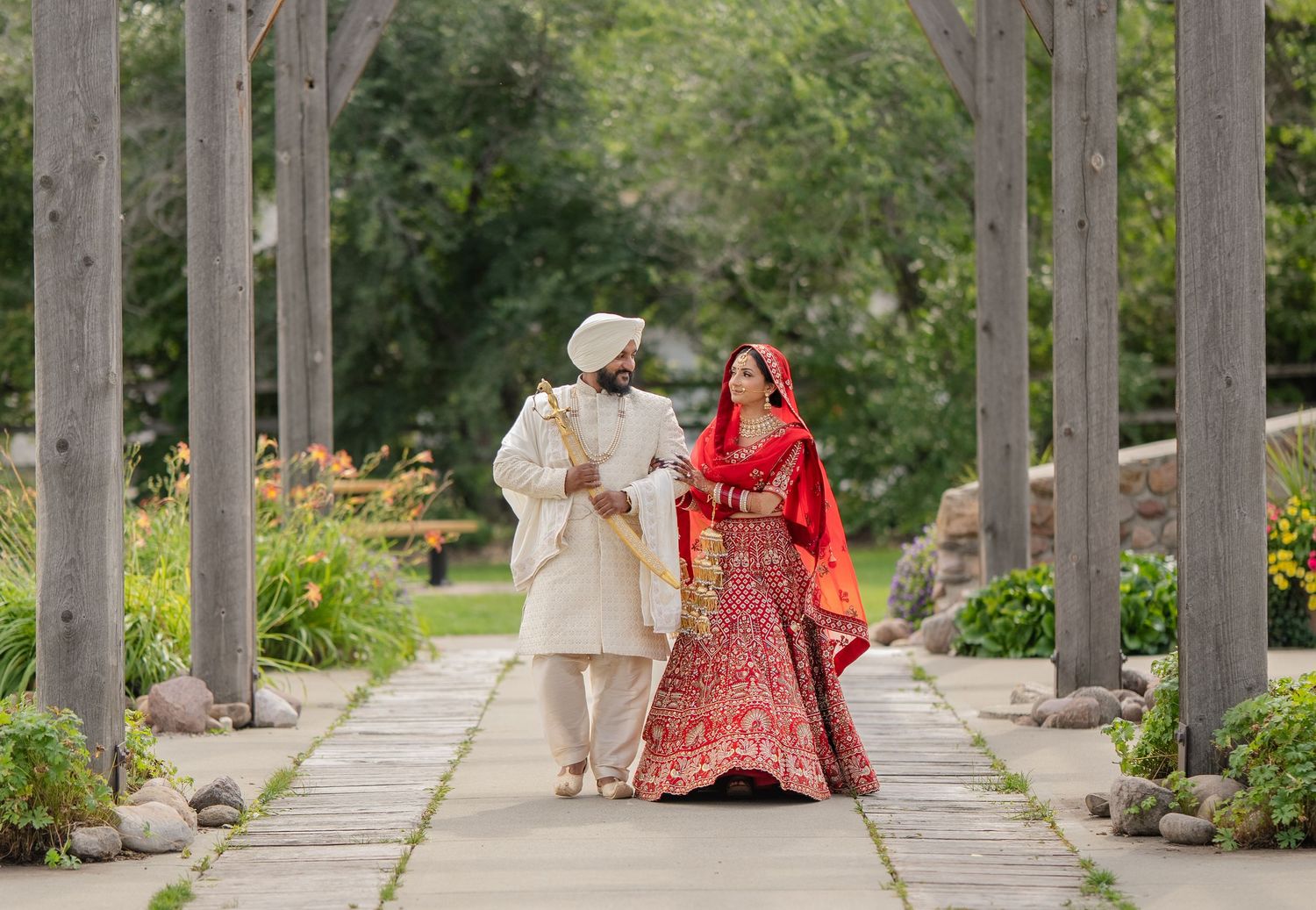 A couple in formal attire walks together through a scenic garden pergola path with stone columns.