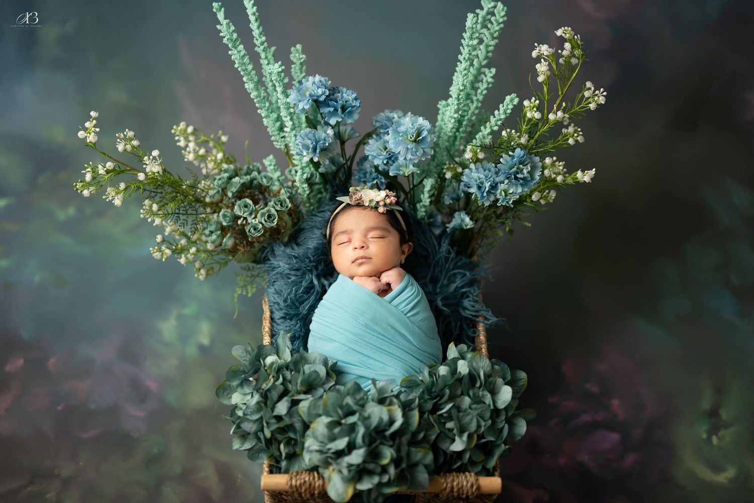 Sleeping newborn swaddled in teal wrap nestled among flowers and greenery in wooden basket.