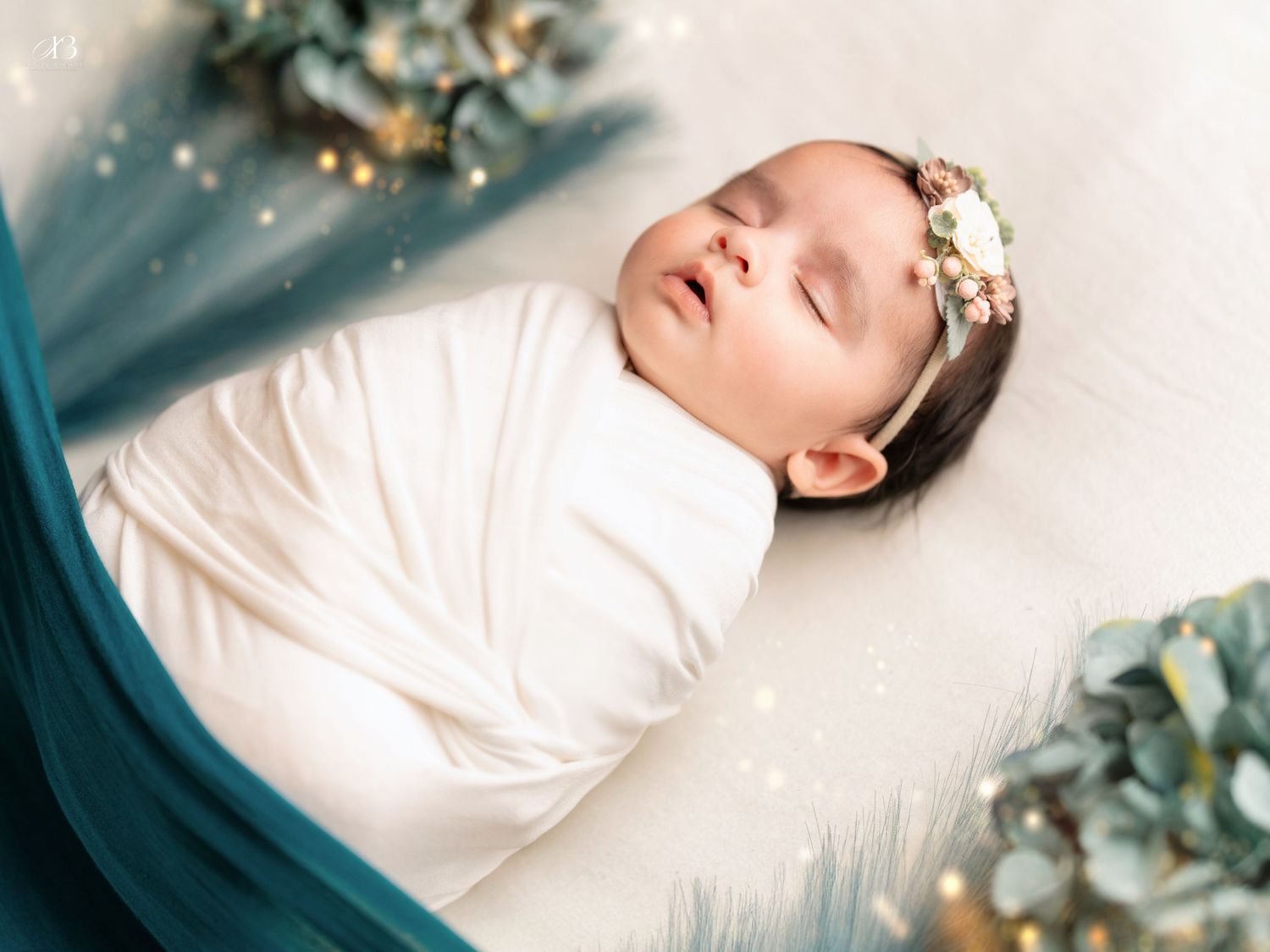 Sleeping newborn wrapped in white with floral crown against dreamy blue backdrop.