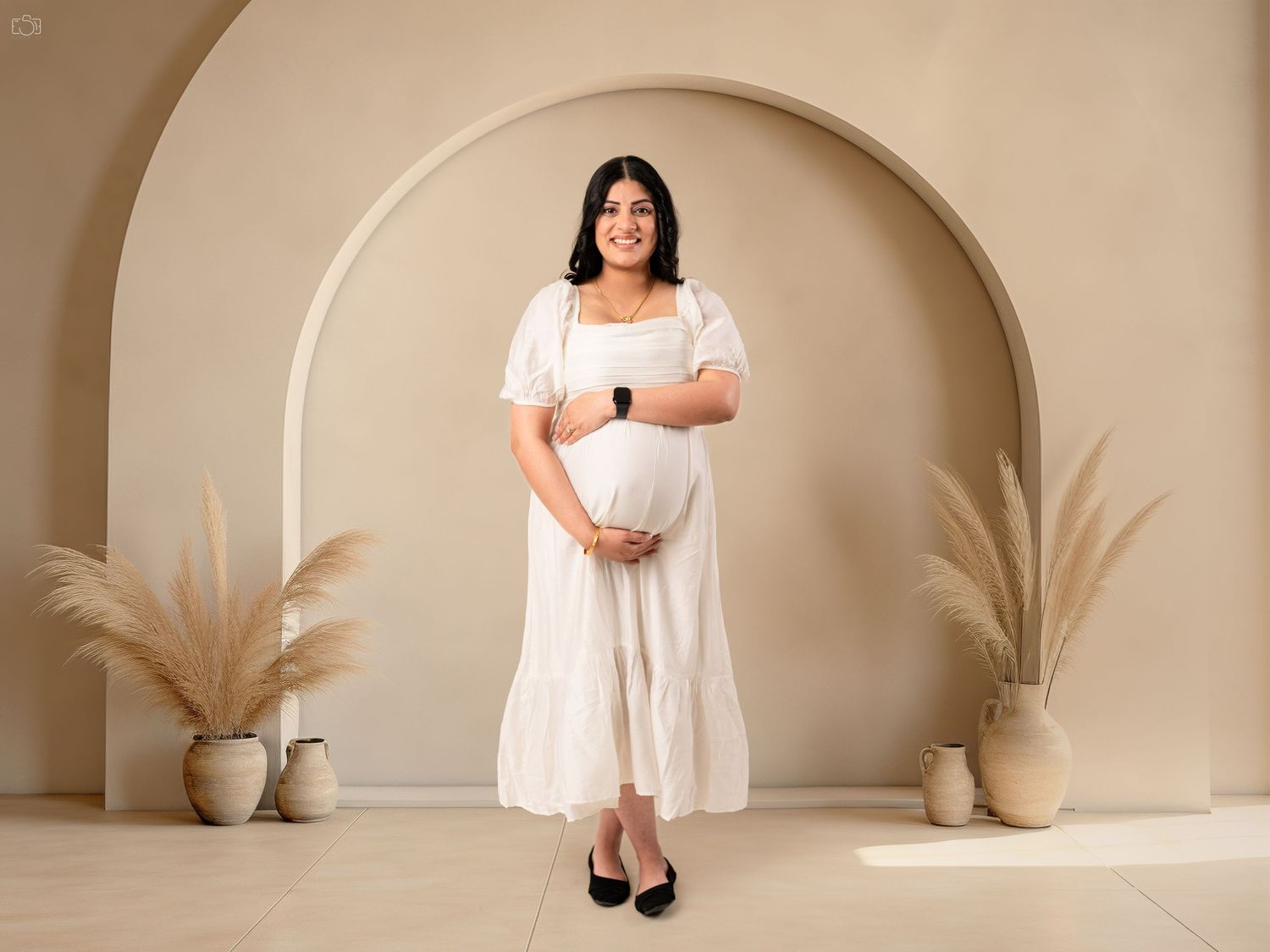 A maternity portrait in a white dress against a neutral archway with pampas grass.