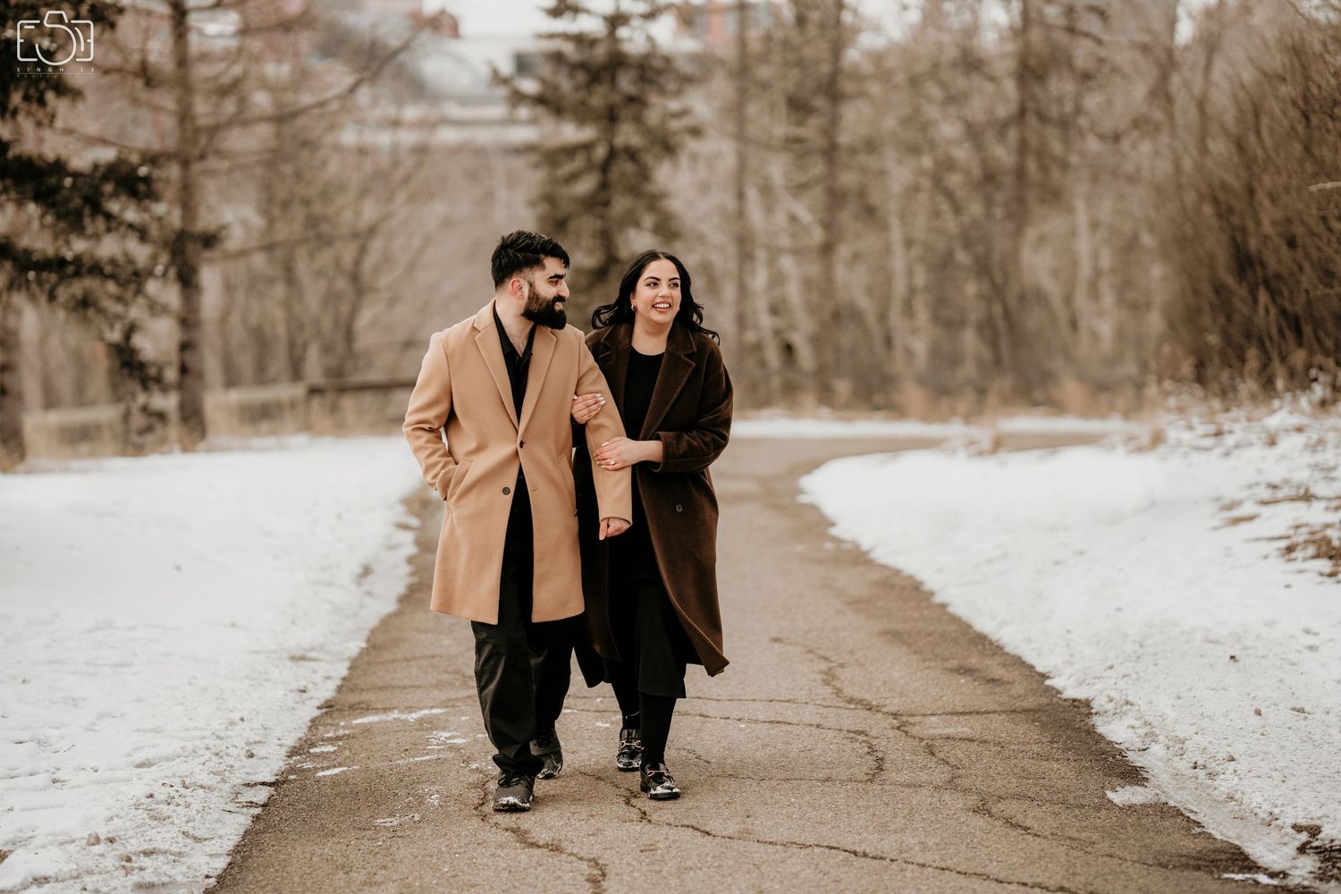 A couple in matching camel coats walking together on a snowy winter path.