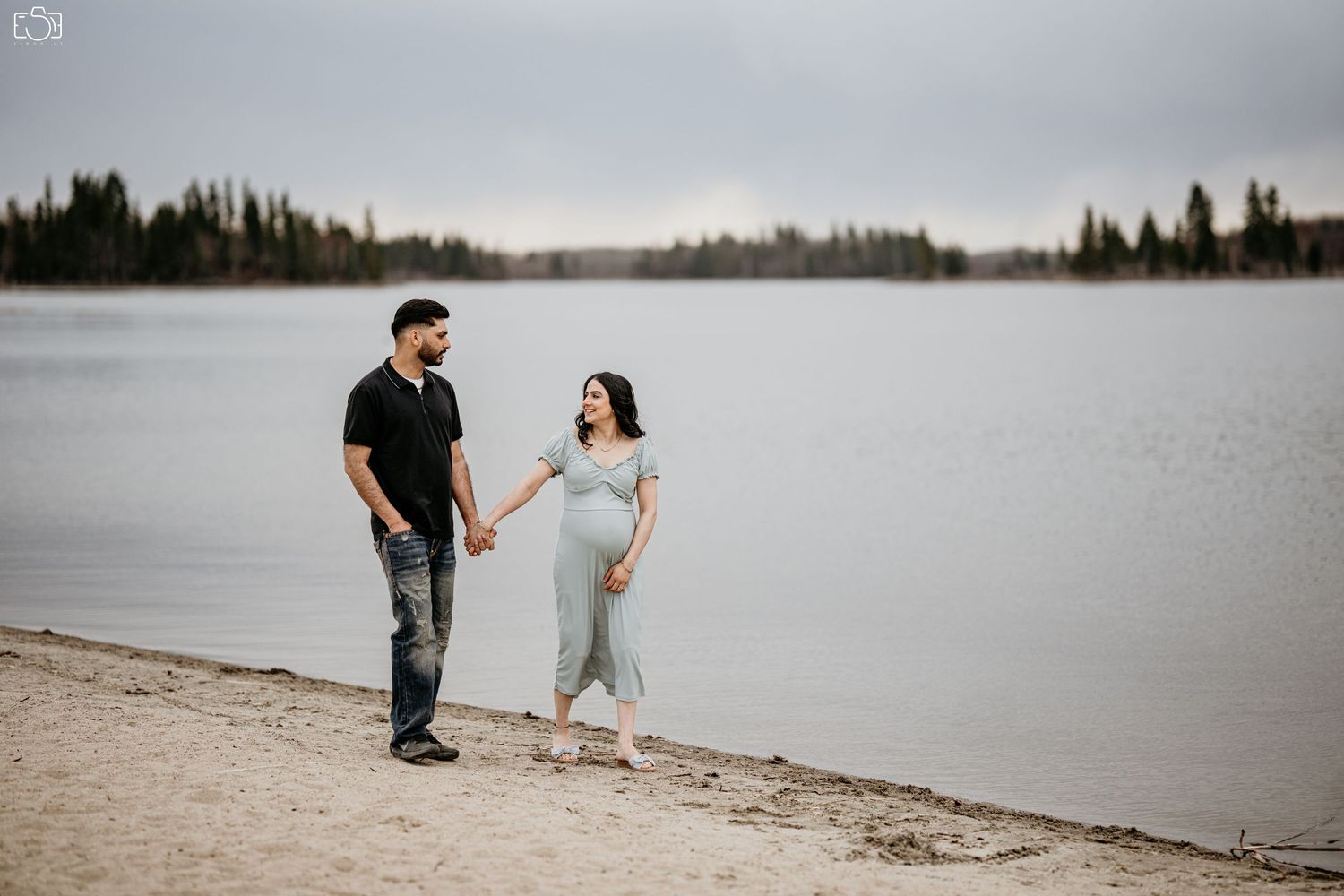 Expectant couple walking along lakeside beach with forest in background.