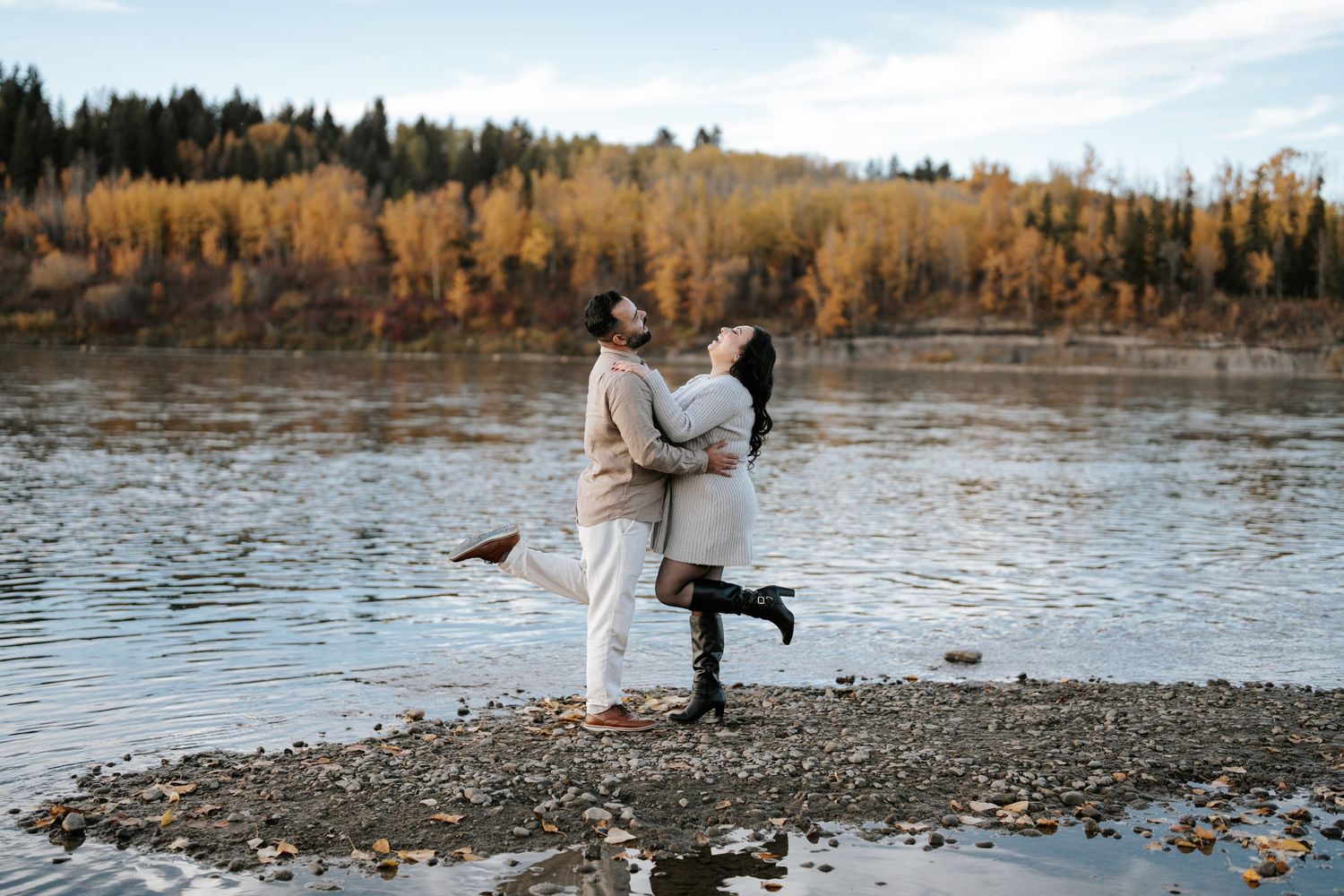 Romantic couple embracing on rocky riverbank with autumn forest in background.