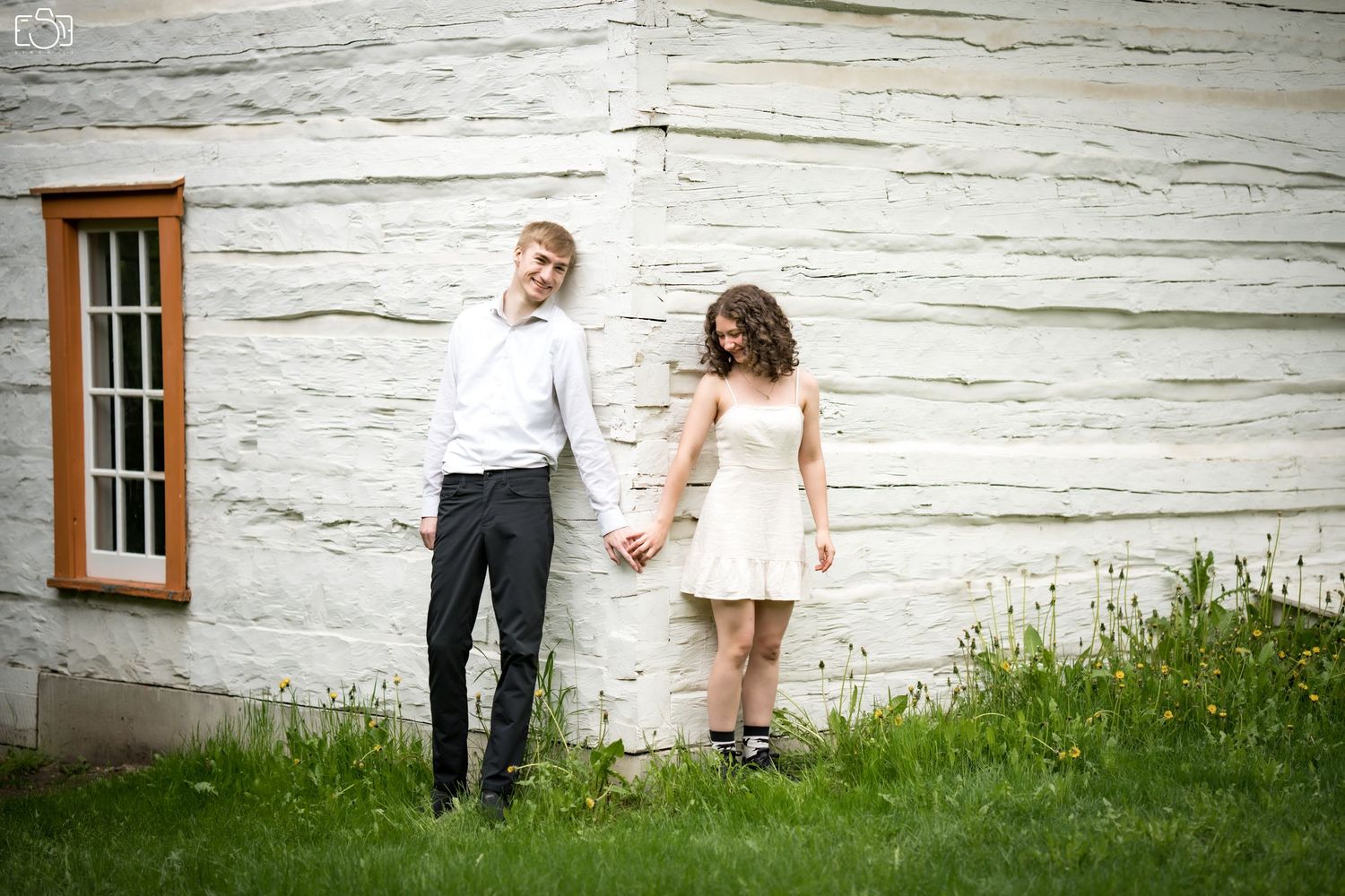 Couple holds hands while standing against white rustic wall in garden setting.