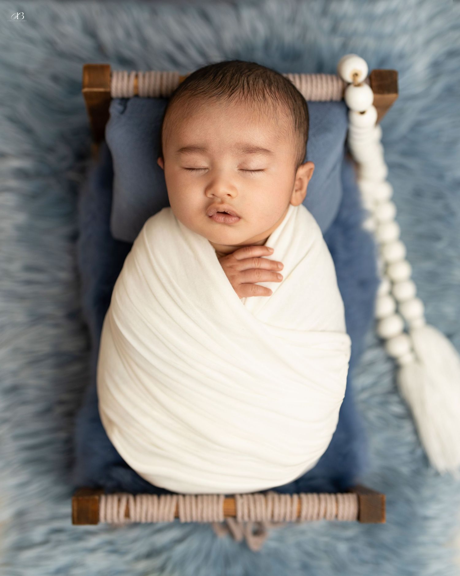 Swaddled baby sleeping on blue miniature bed with white beaded decoration.