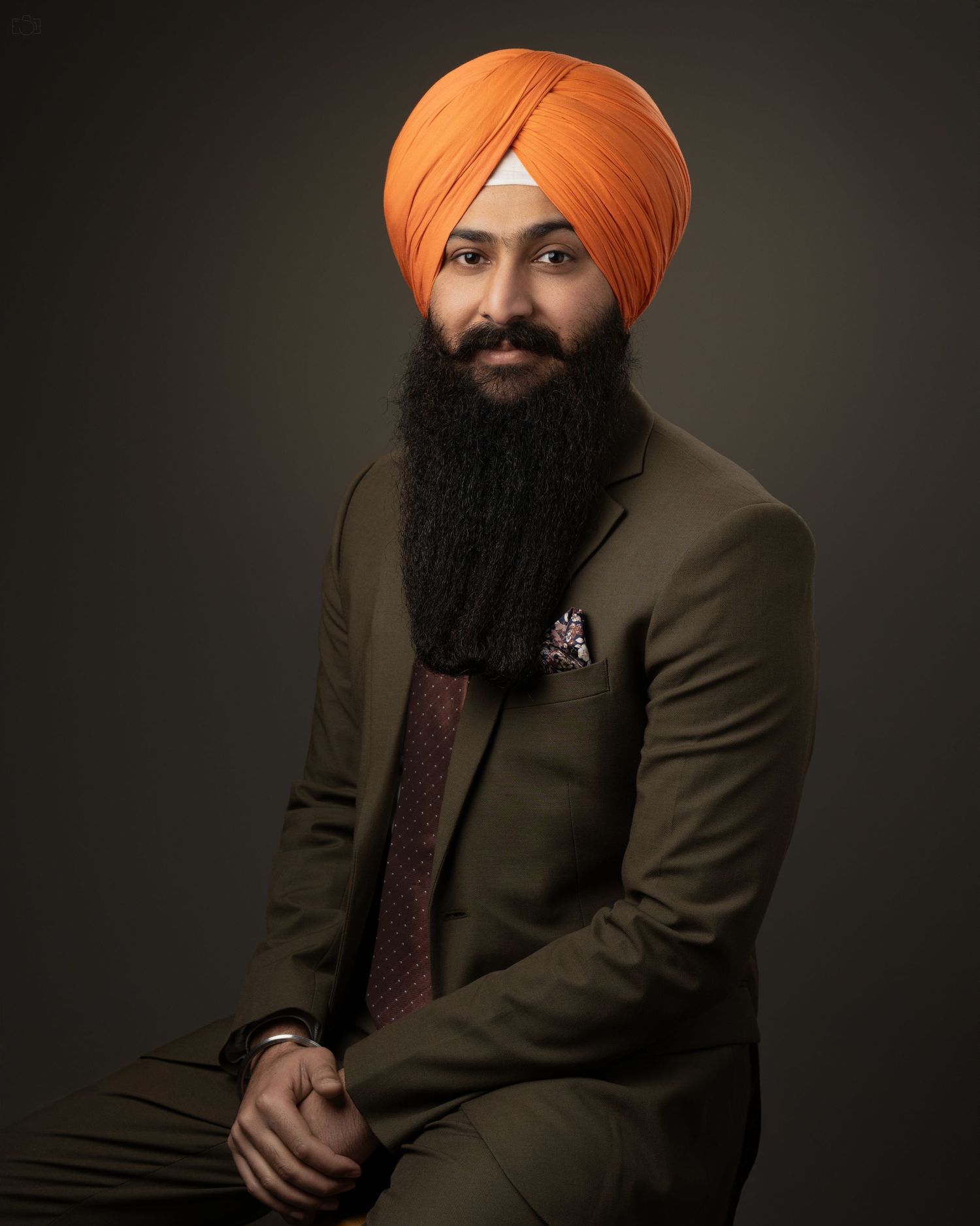 Professional headshot of a person in an olive suit jacket and orange turban against a dark background.