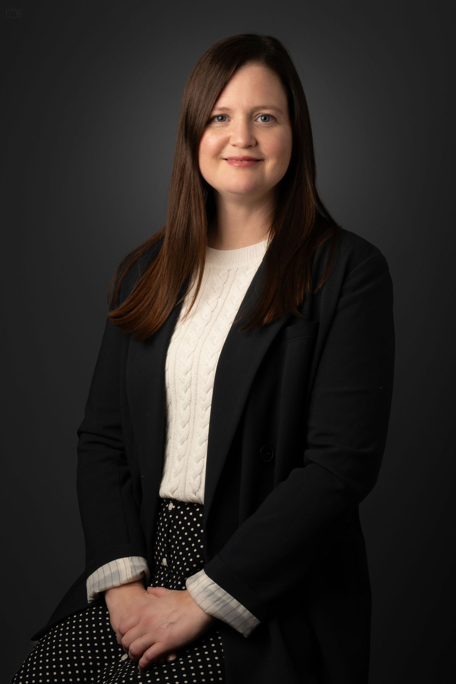 Professional headshot of a person wearing a black blazer and white cable knit sweater.