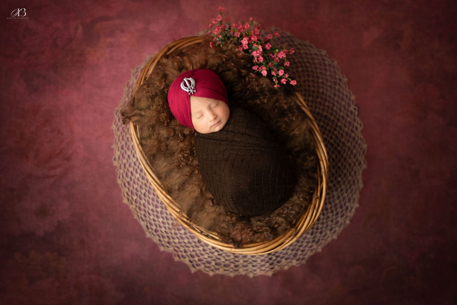 Sleeping newborn in dark wrap nestled in round basket with red floral accent.