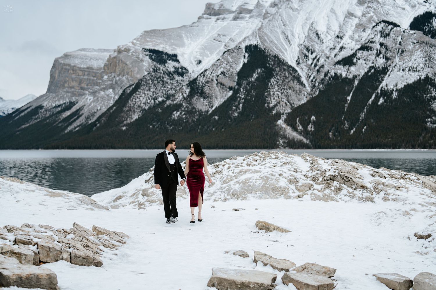 Couple standing on snowy rocks with majestic mountain peaks and lake in the background.