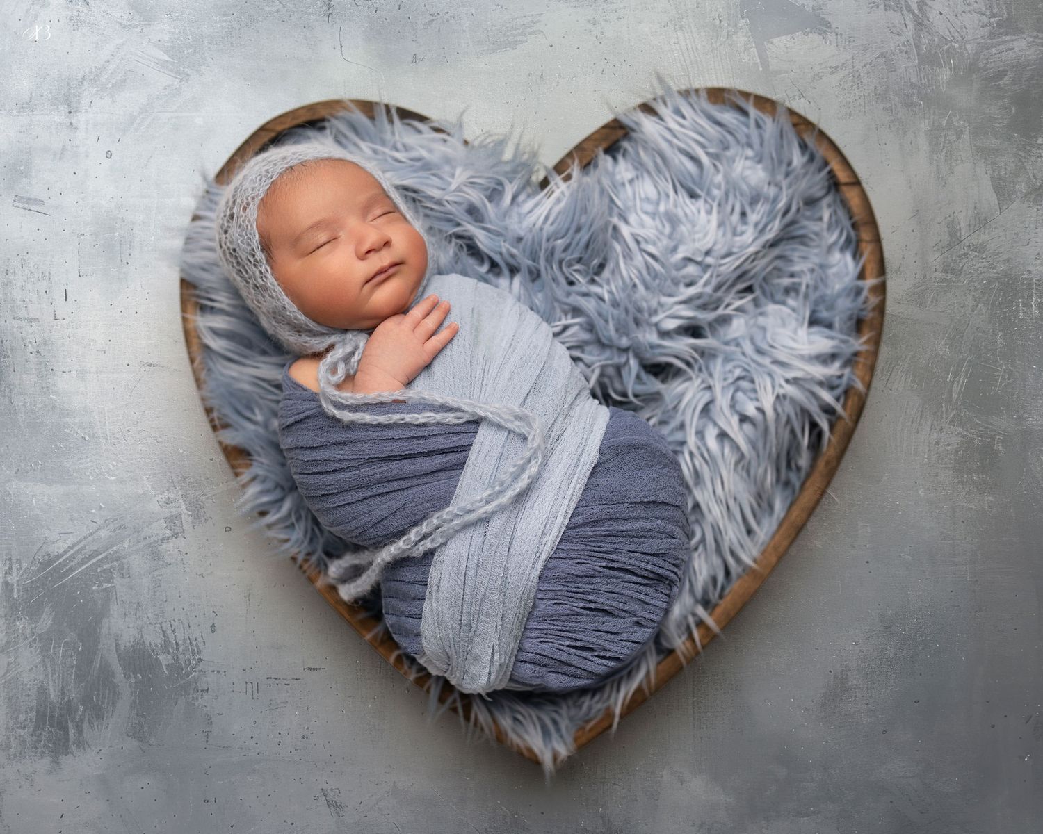 Peaceful newborn wrapped in grey fabric lying in heart-shaped container with fluffy bedding.