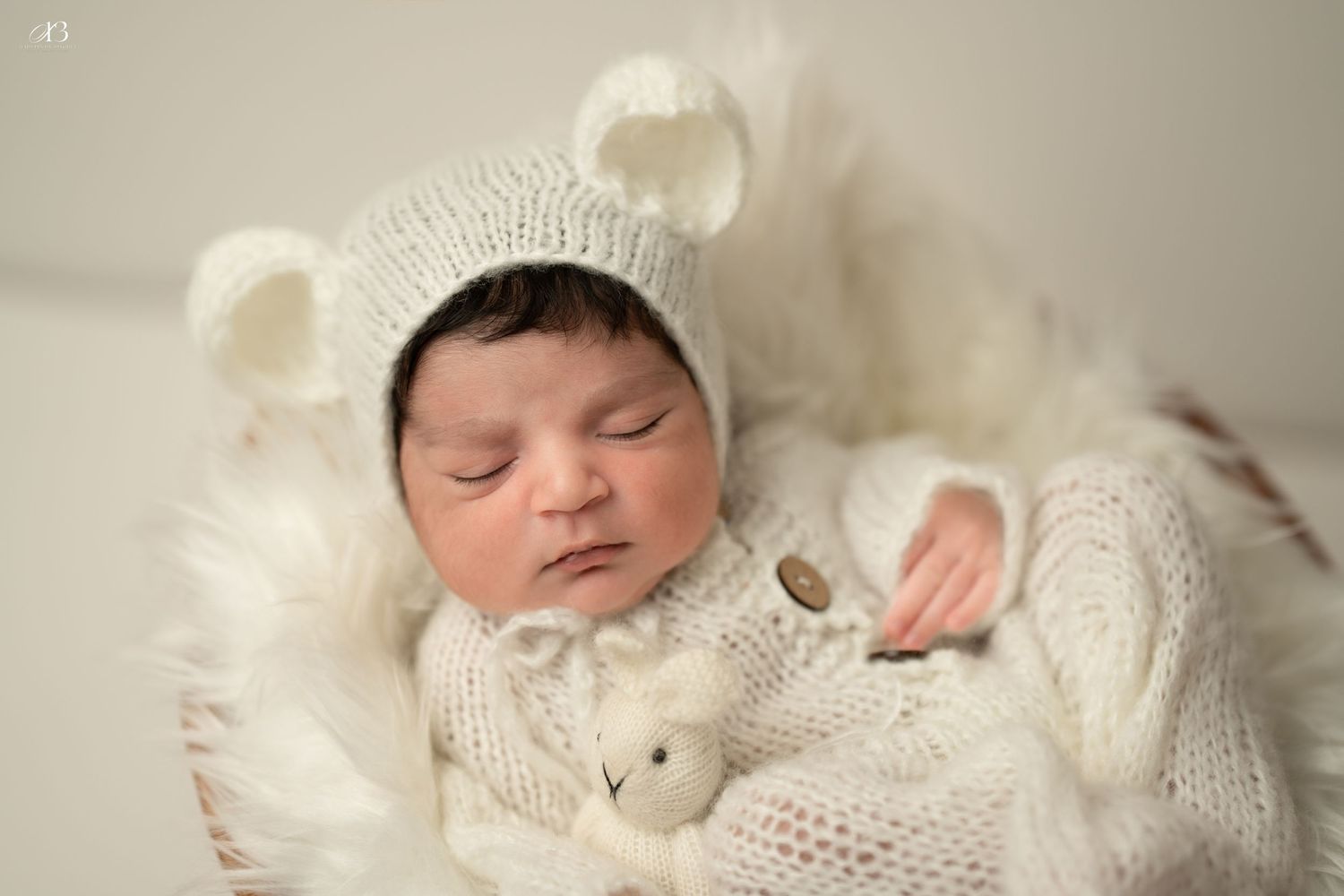Sleeping newborn in white knit bear outfit cuddling plush bunny toy.