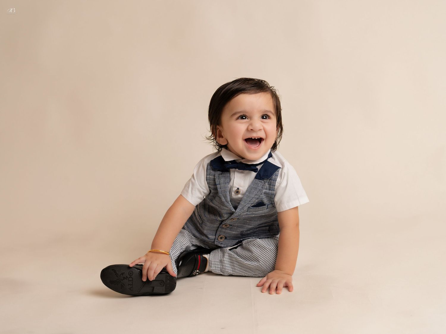 Baby boy in formal vest and bowtie outfit sits smiling against beige backdrop.