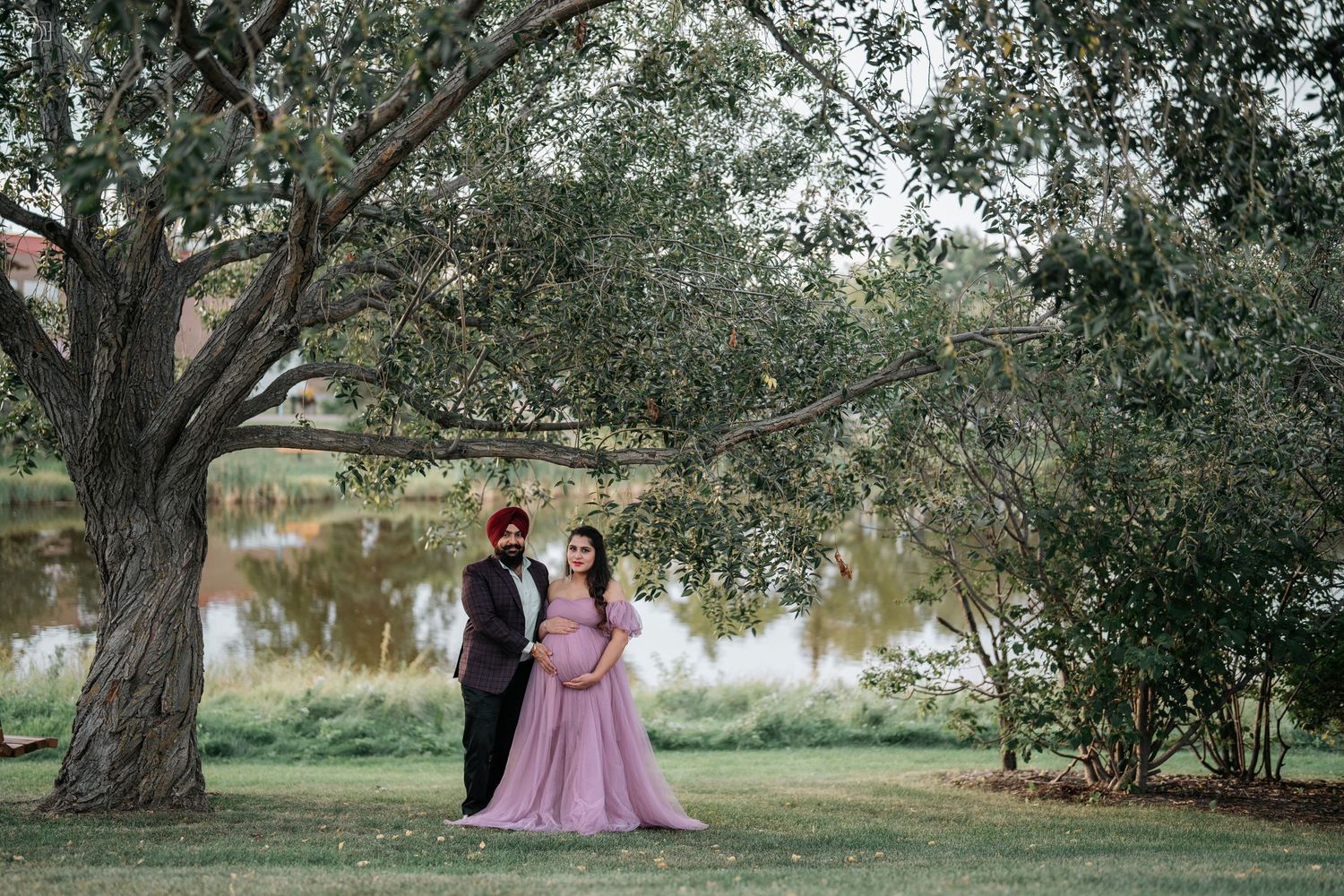 A romantic couple poses by a lake under a large tree at sunset.