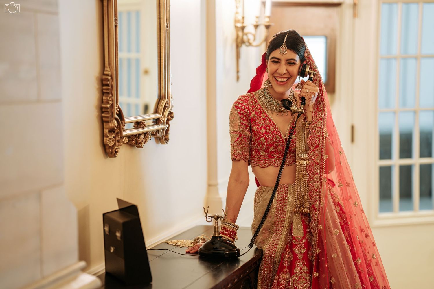 Bride in traditional red lehenga poses elegantly by ornate mirror during wedding preparations.