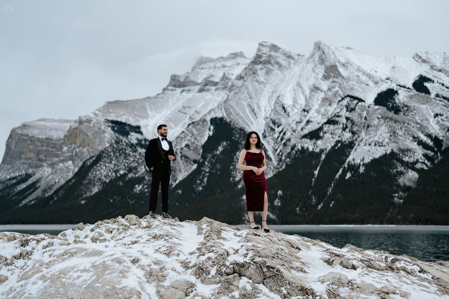 Couple poses dramatically on mountain cliff edge with snow-capped peaks behind them.