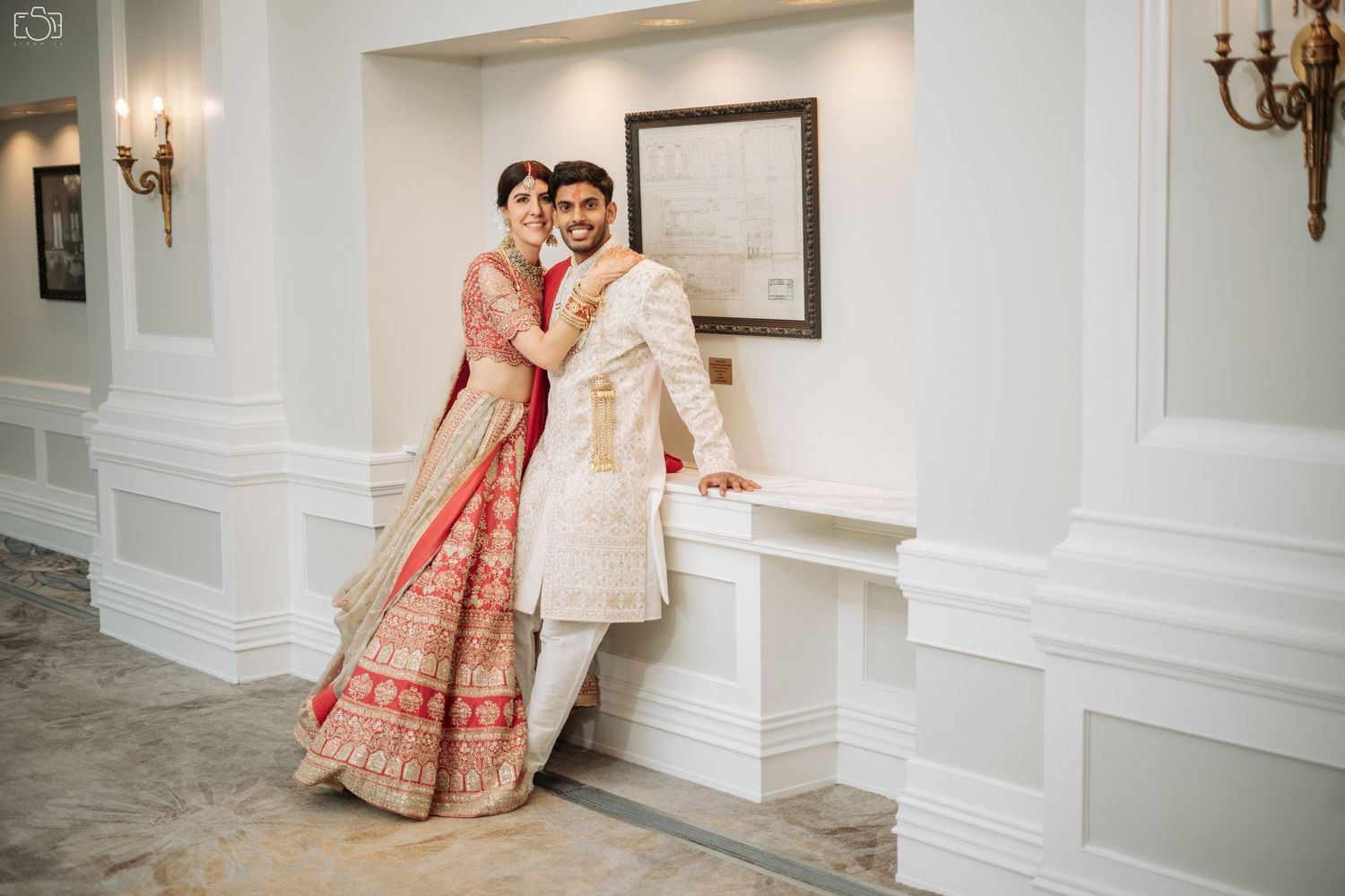 A wedding couple in traditional Indian attire posing in an elegant white interior hallway.