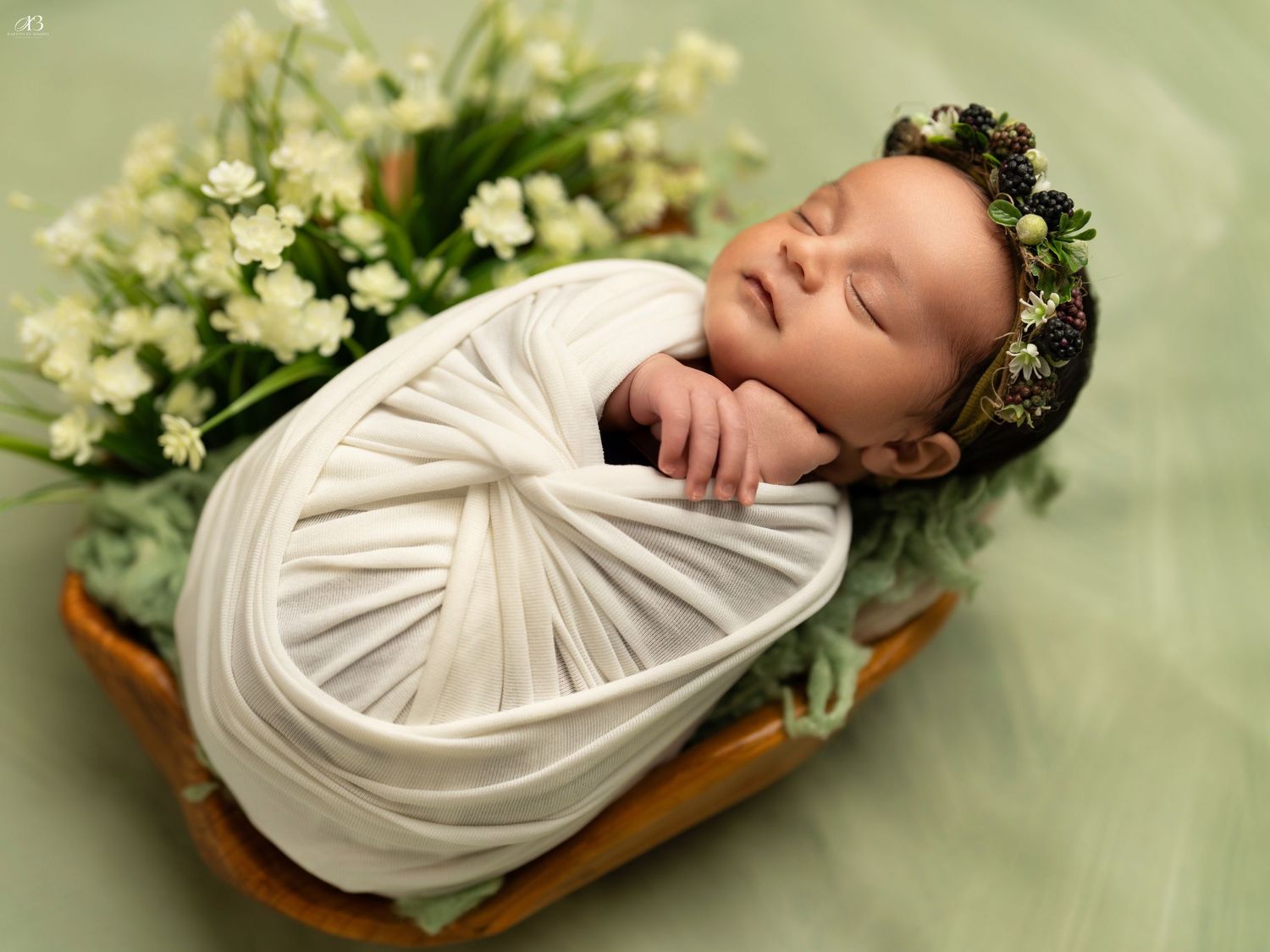 Newborn in white wrap and floral crown sleeps in wooden basket with spring flowers.