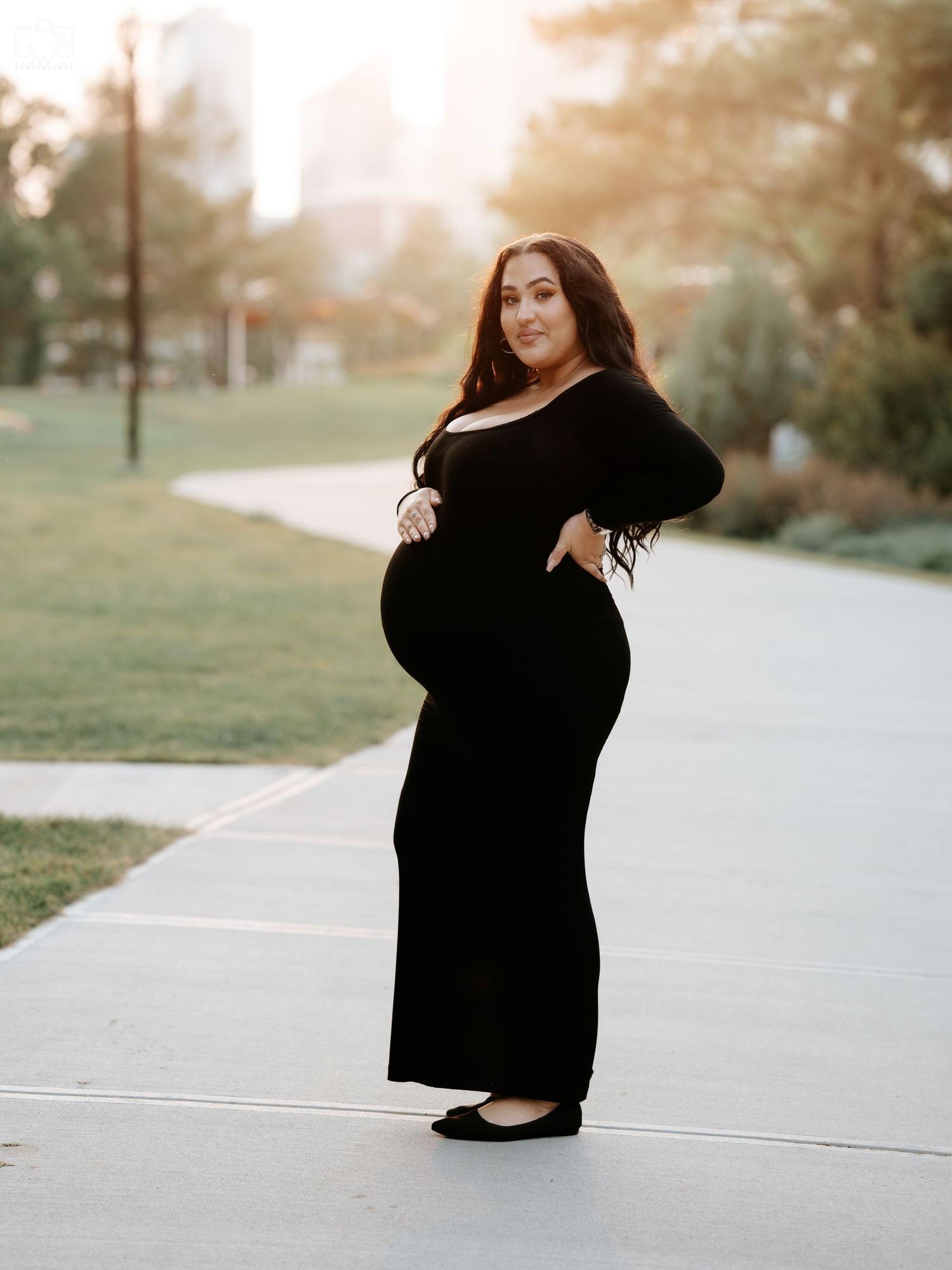 A woman in a black maxi dress poses on a sidewalk during golden hour in an urban park.