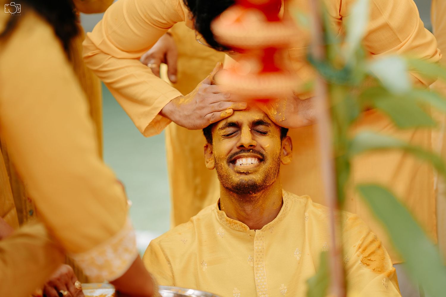 Traditional pre-wedding ritual with hands placing turmeric on seated person in yellow attire.
