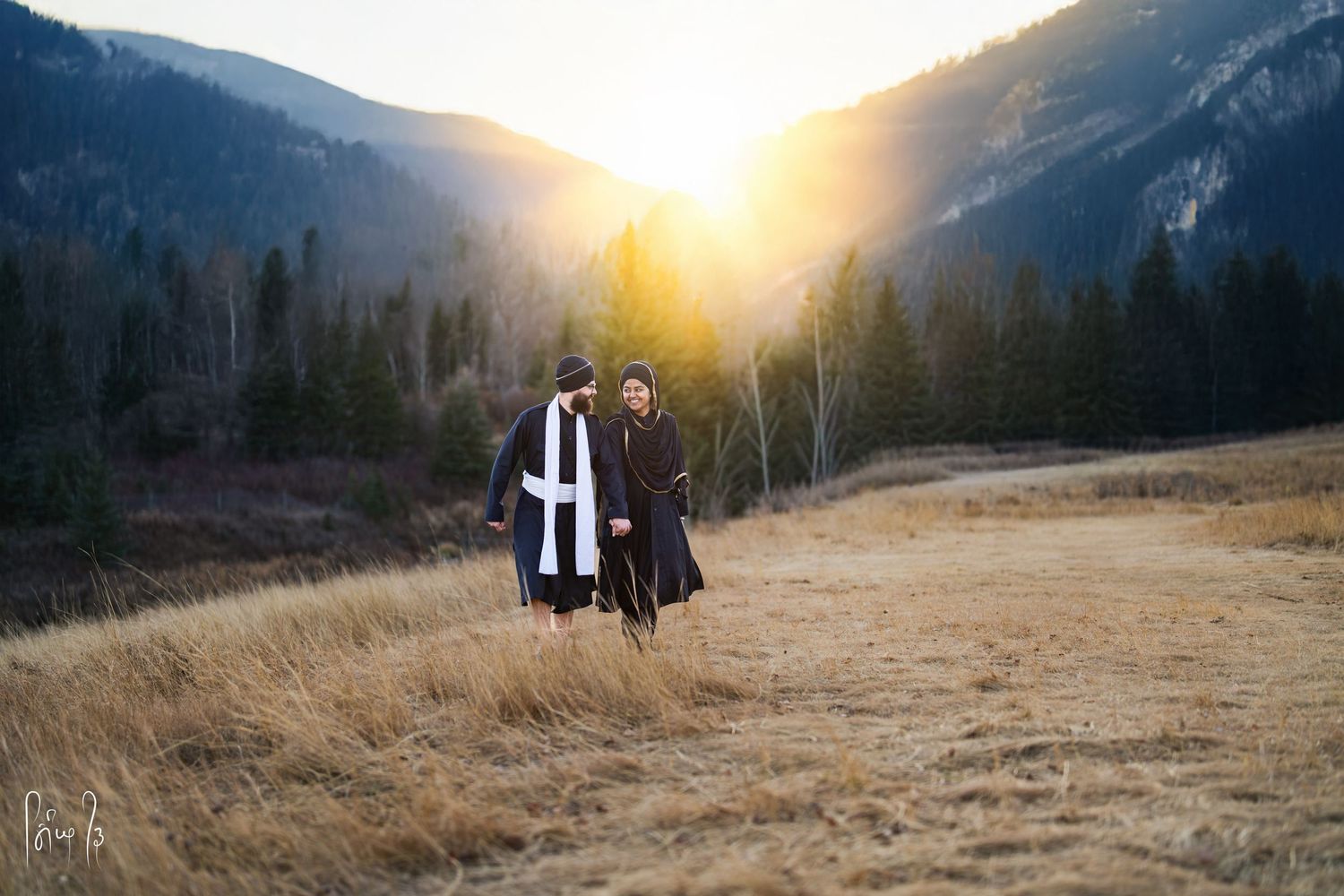 Two people stand together on a golden mountainside meadow at sunset with evergreen forest in the background.