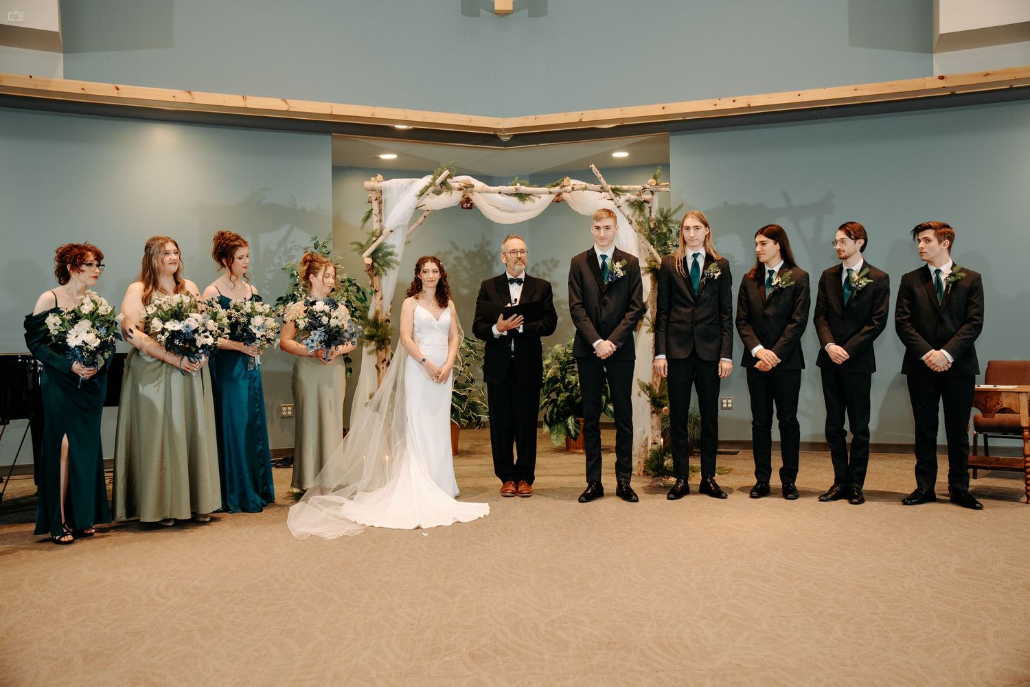 A wedding party lined up against a blue wall under an arch with bridesmaids in grey dresses.