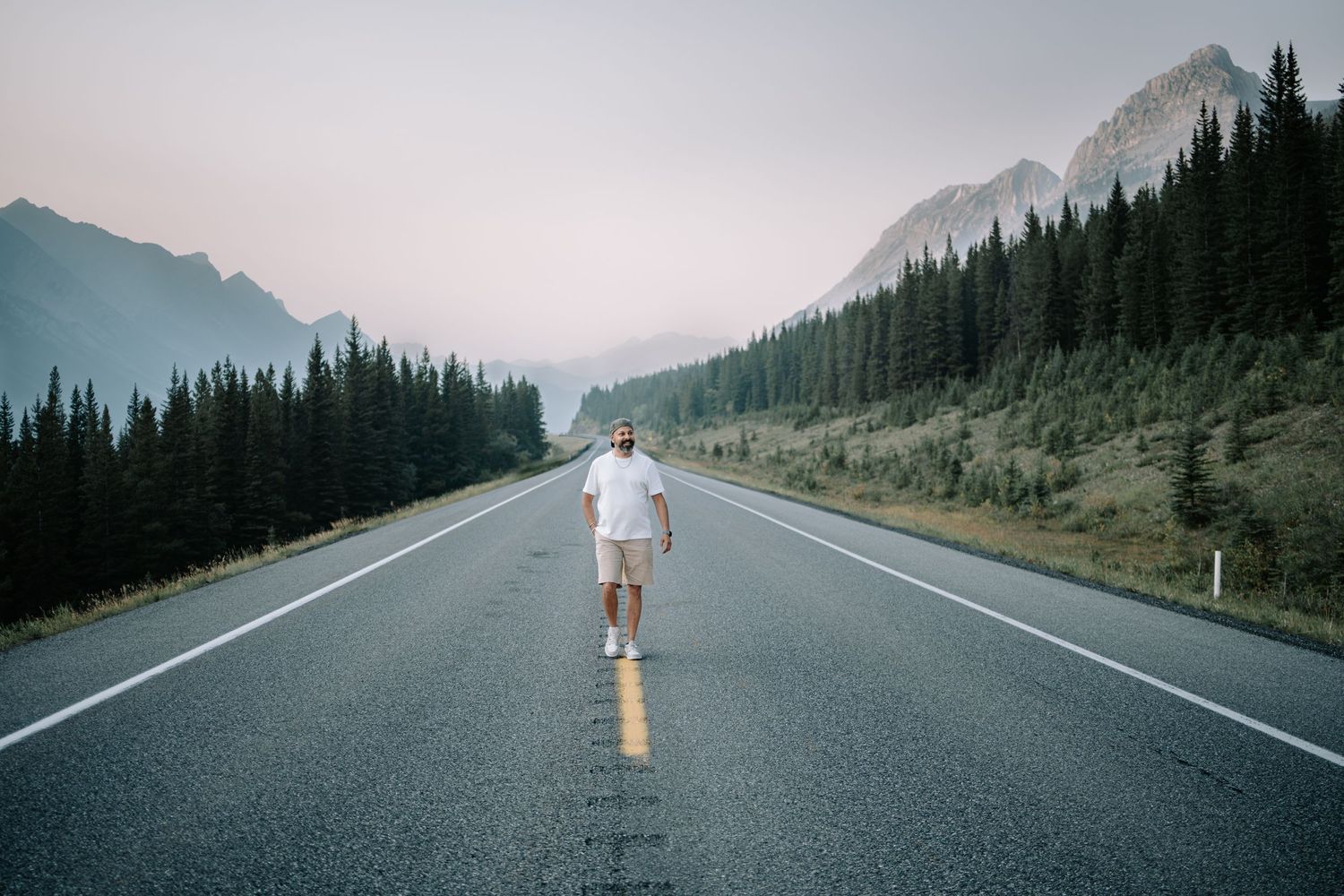 A lone figure stands in the middle of a mountain highway surrounded by pine forests and misty peaks.