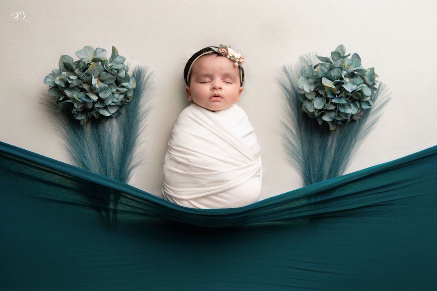 Newborn centered between teal fabric and blue hydrangeas in artistic photography setup.